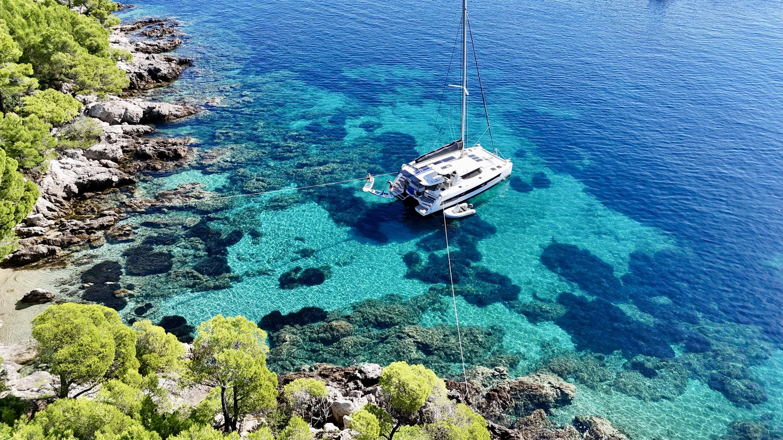 Aerial view of a white luxury yacht anchored near a rocky, tree-lined shoreline with clear turquoise water.