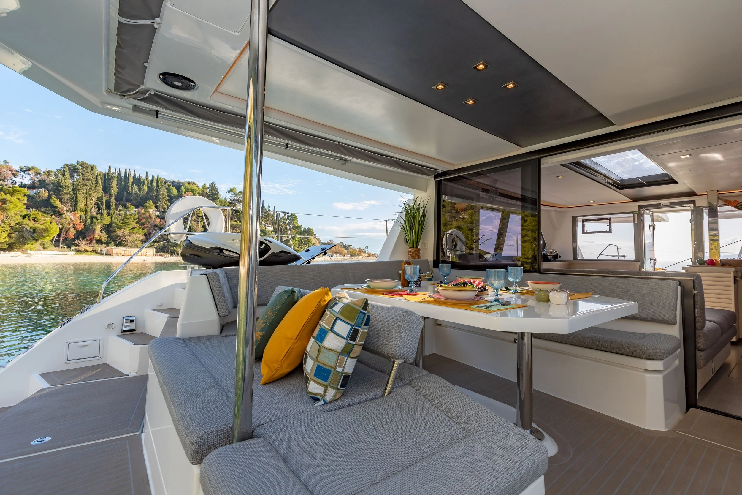Interior of a luxury yacht with a dining table set for a meal, cushions on the seating, and a view of the water and trees outside.