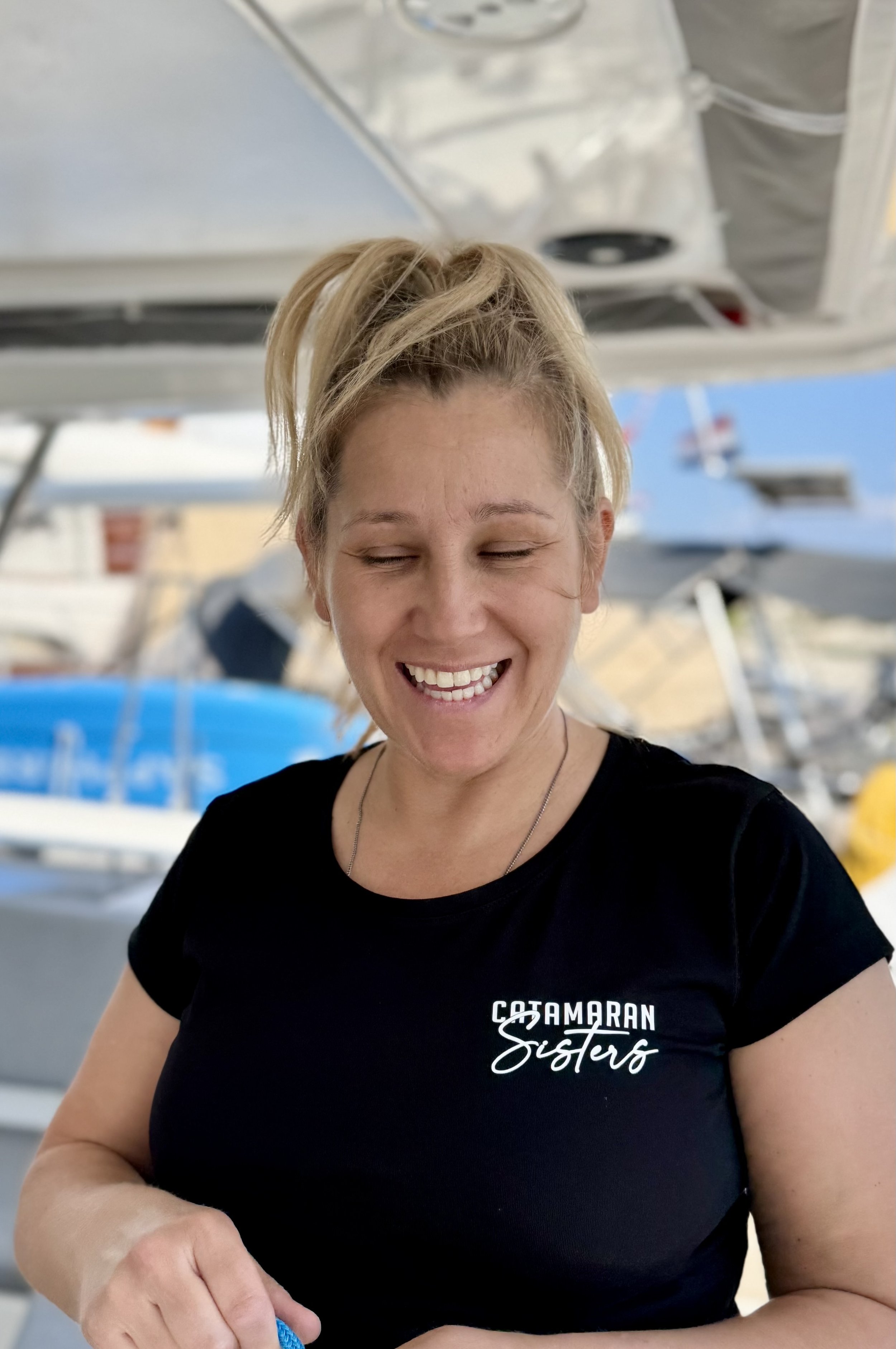 A woman smiling with closed eyes, wearing a black T-shirt that says "COTAMARAN Sisters" and standing near boats.