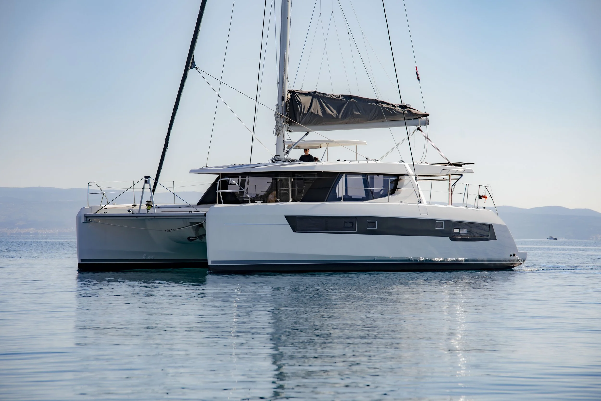 A modern white sailing catamaran yacht with a man on deck, floating on calm water with distant hills in the background and a clear sky.