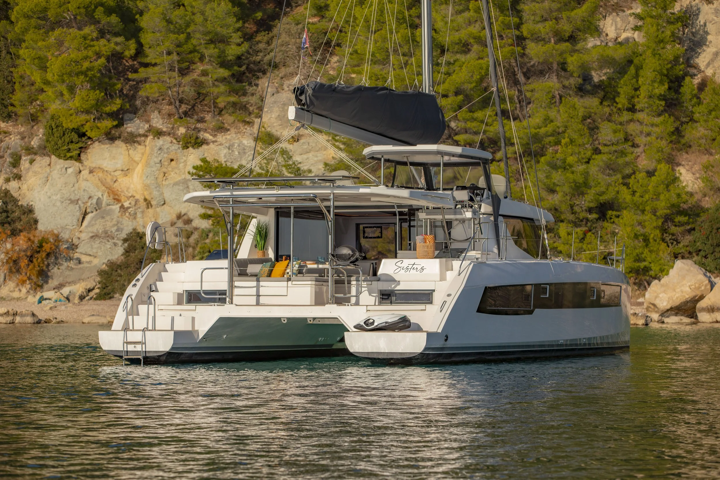 A modern white yacht with a spacious deck is anchored near a rocky shoreline with green pine trees.