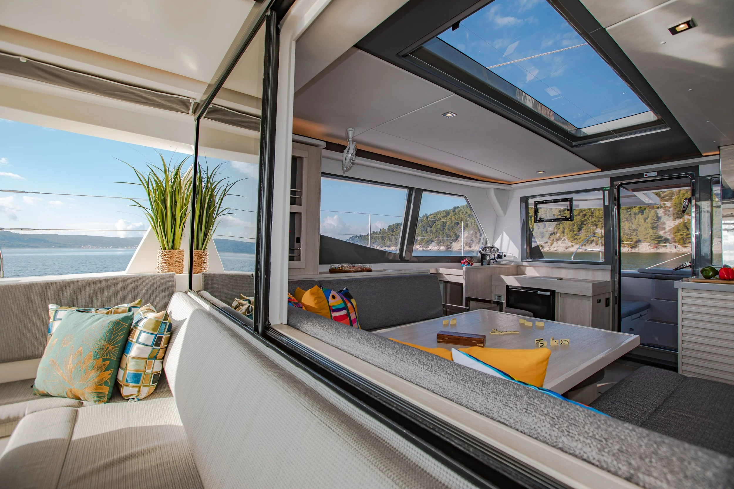 Interior of a yacht with a dining area, sunlight coming through windows and skylight, patterned pillows on beige sofa, board game pieces on table, view of water and shoreline outside.