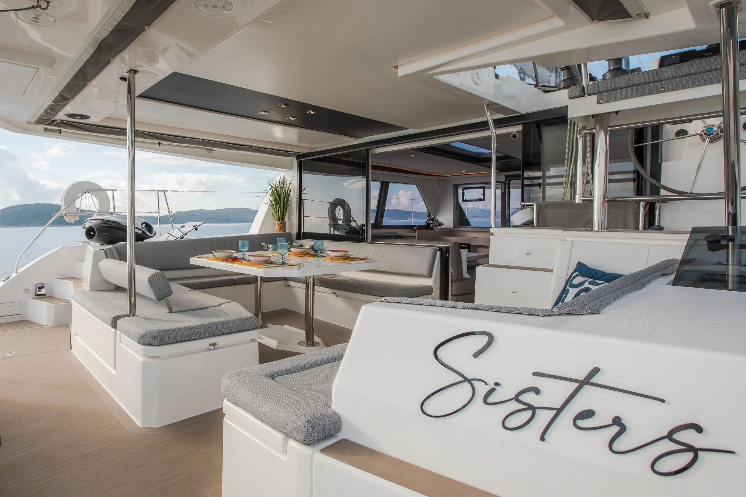 The deck of a yacht with a dining area set with bowls, glasses, and plates, surrounded by cushioned bench seating, overlooking water and distant islands, with indoor living space visible through glass sliding doors.