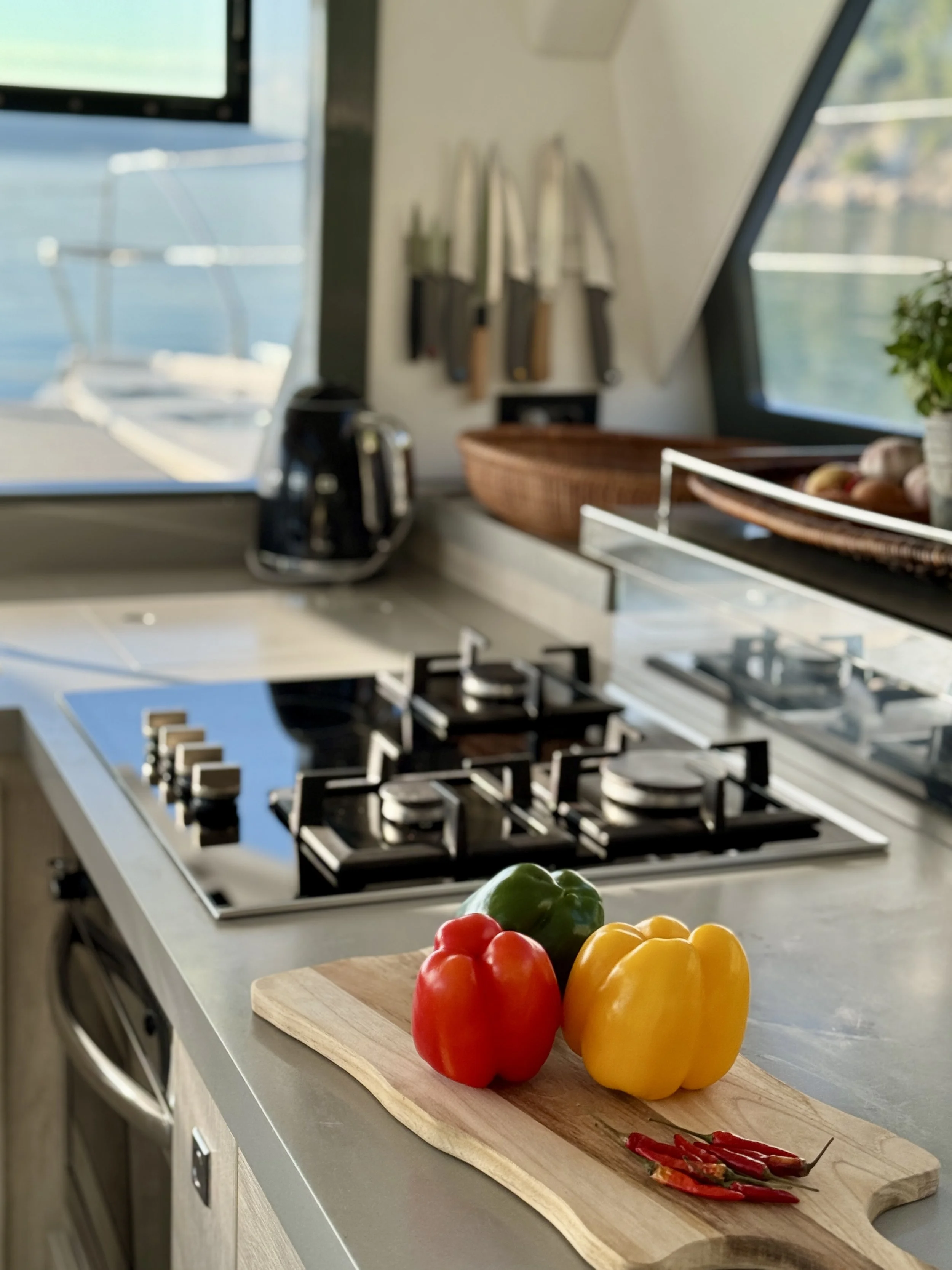 Colorful bell peppers and red chili peppers on a wooden cutting board on a kitchen counter with a stove, coffee maker, and knives in the background, near a window with a view of water.