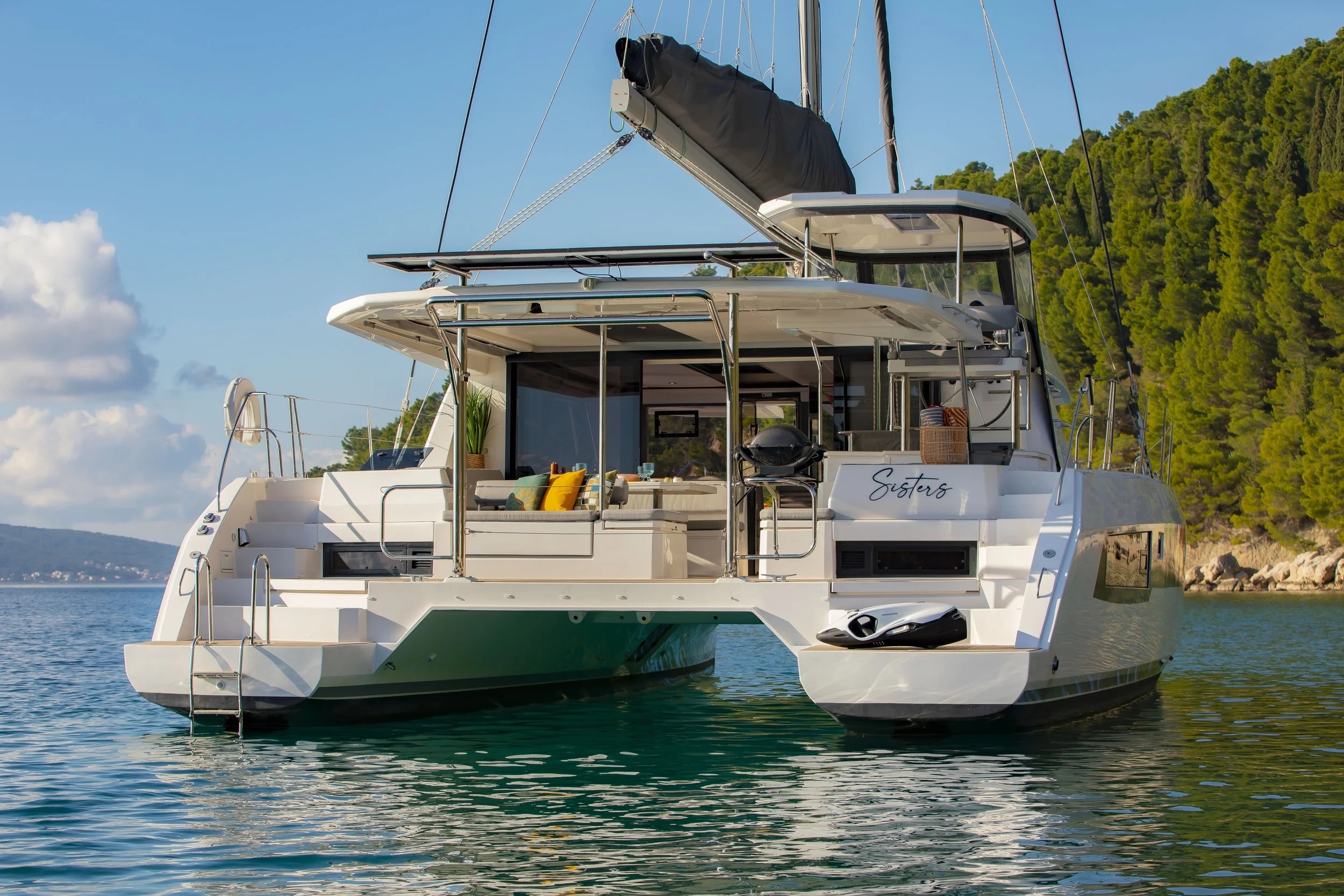 A white yacht named 'Sisters' on calm water near a forested shoreline, with a seating area and various accessories on deck.