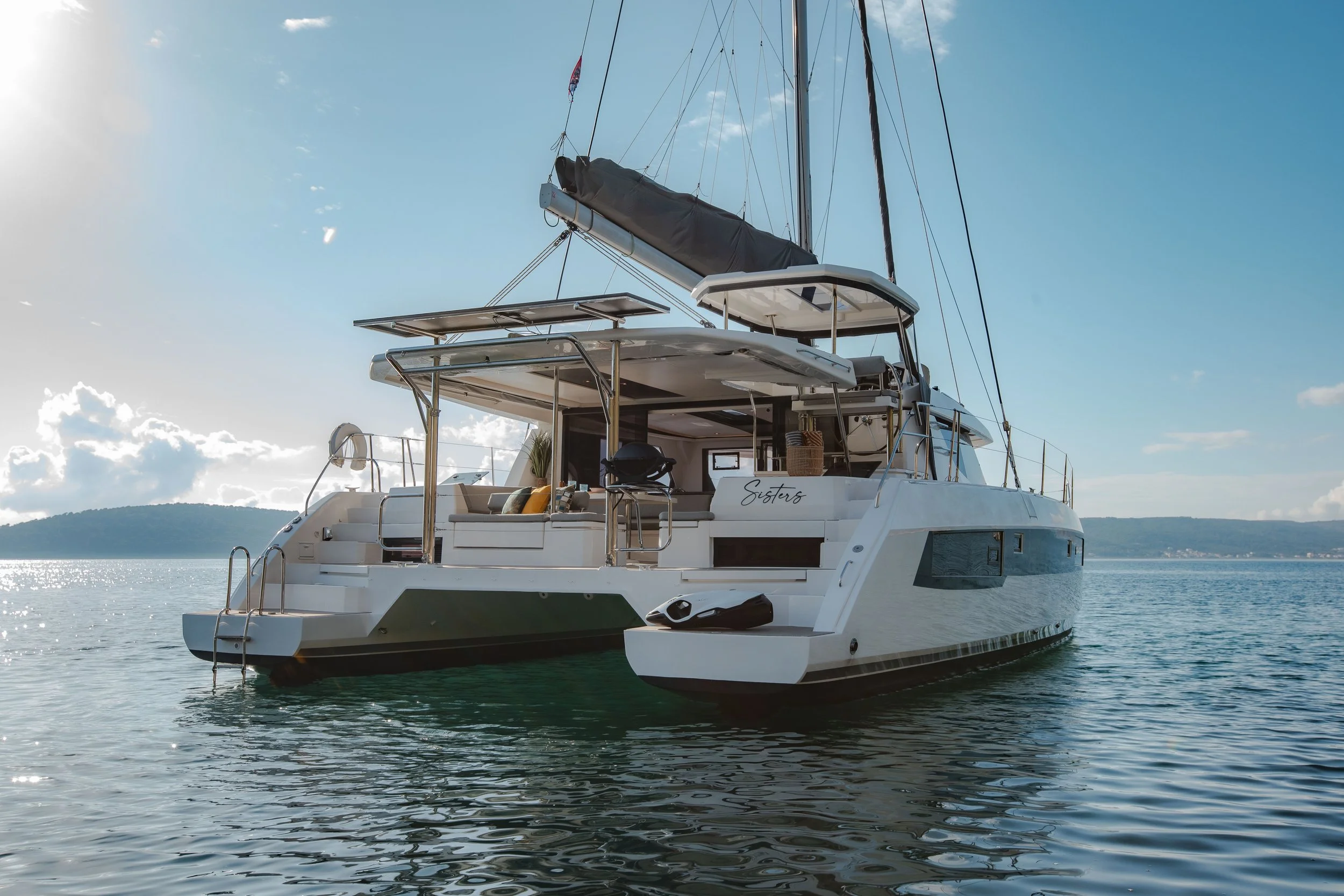 A modern white sailboat with a black sail cover floating on calm water with a hilly island in the distance and a partly cloudy blue sky.