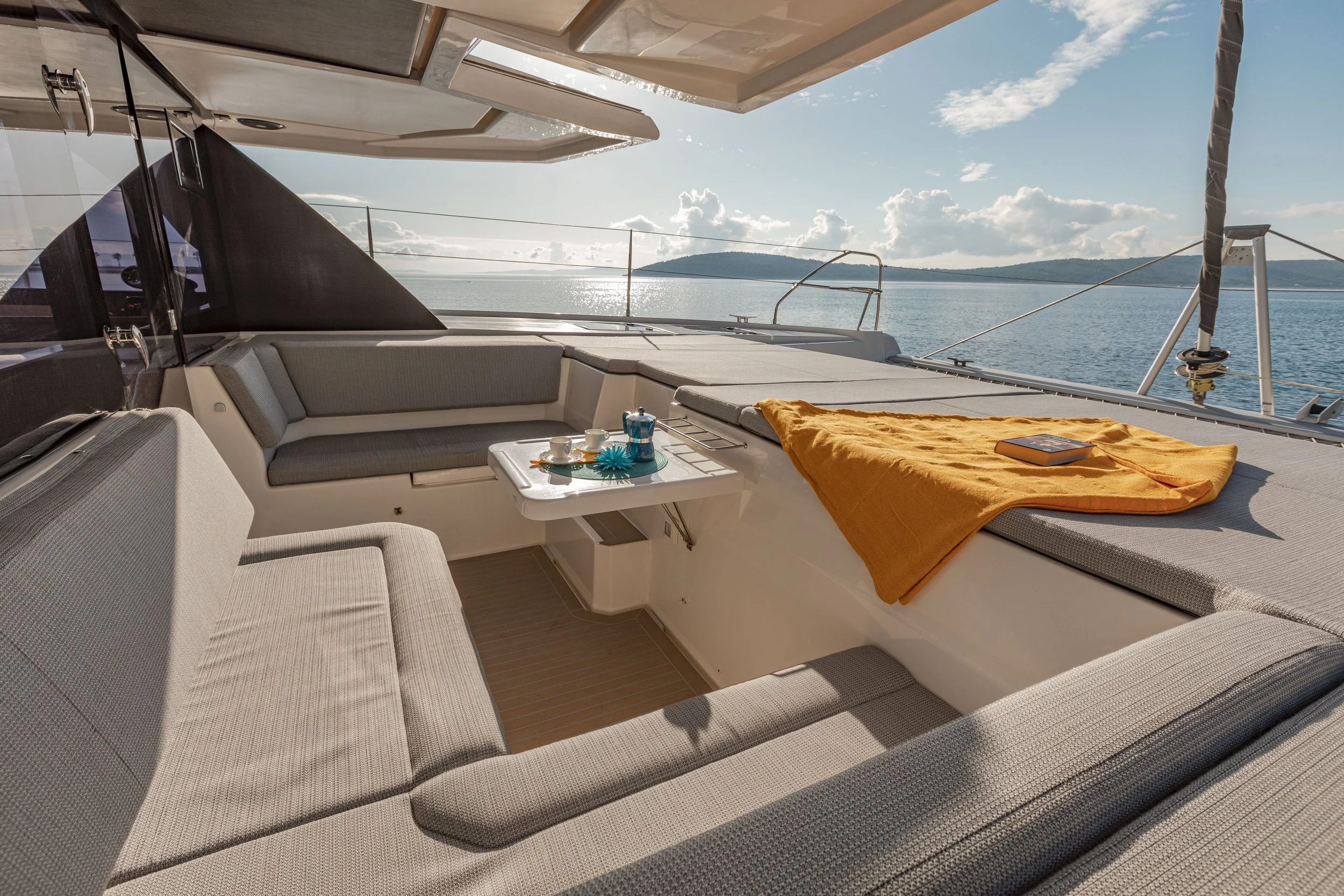 View of a yacht deck with seating area, a small table set with cups and a teapot, a rolled-up orange towel, and a book, overlooking calm water and a distant land under a partly cloudy sky.