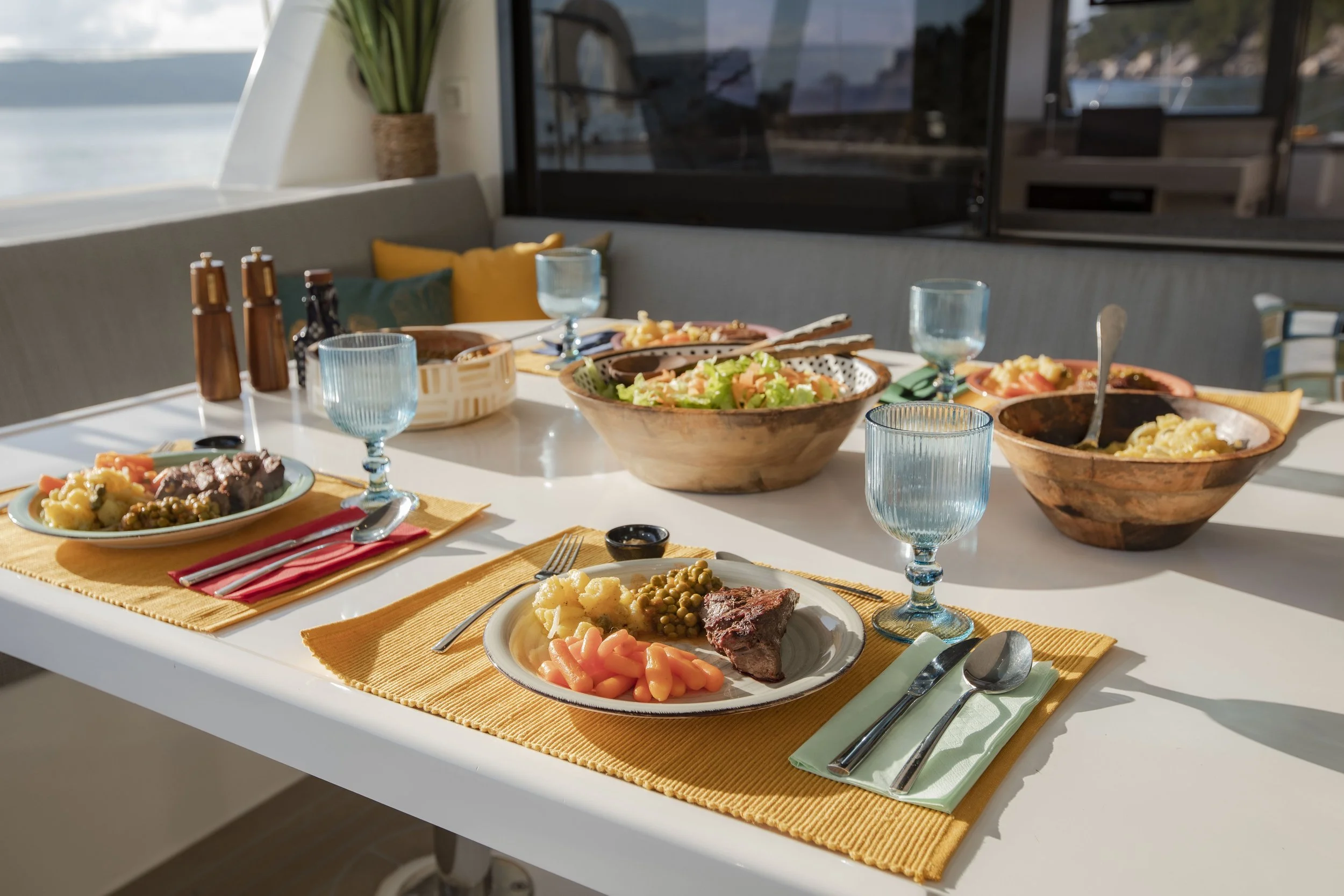 A dining table set with plates of food, salad bowls, and drinking glasses, overlooking a waterfront view during daylight.