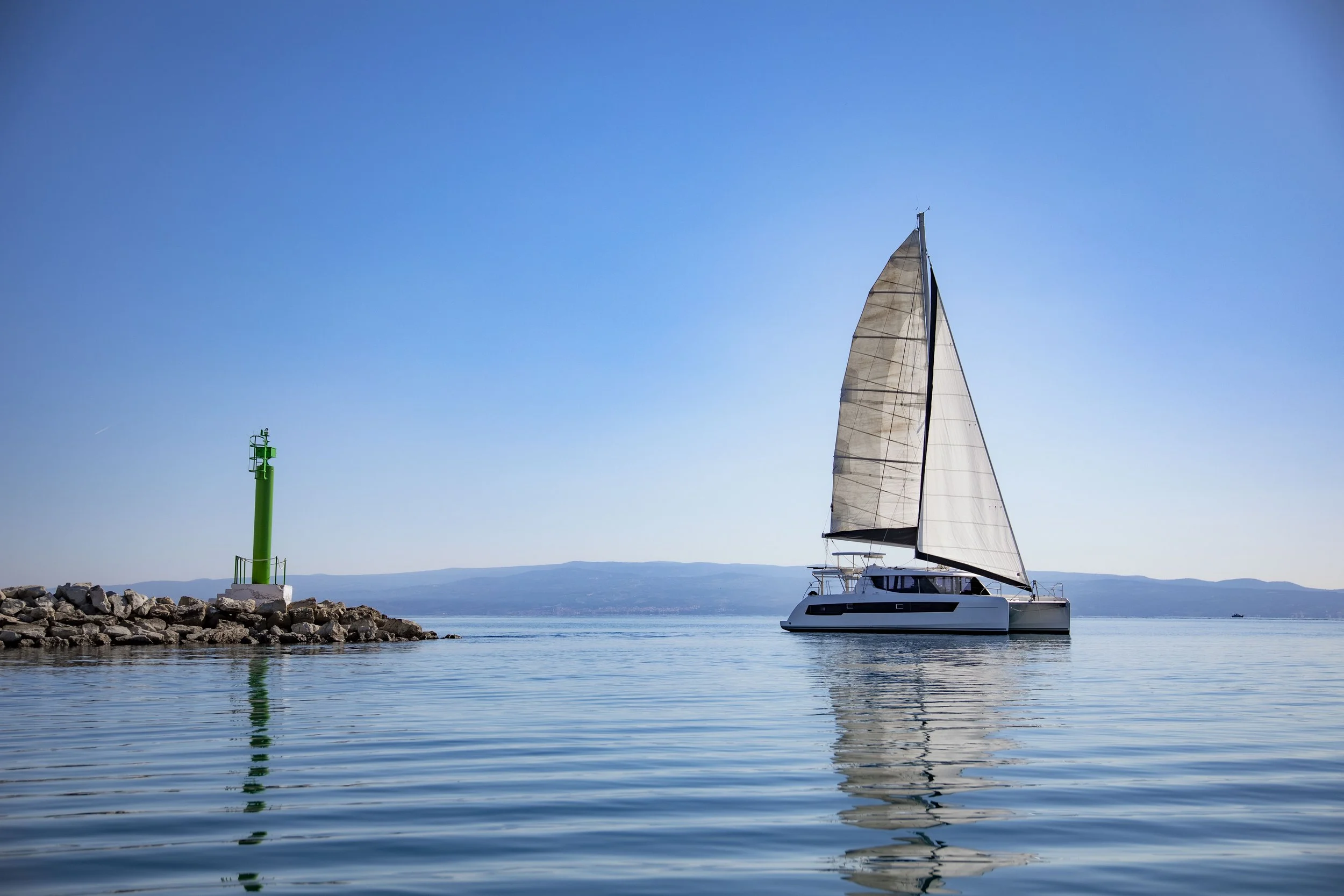 A sailboat on calm water near a rocky pier with a green lighthouse, and distant hills in the background under a clear blue sky.