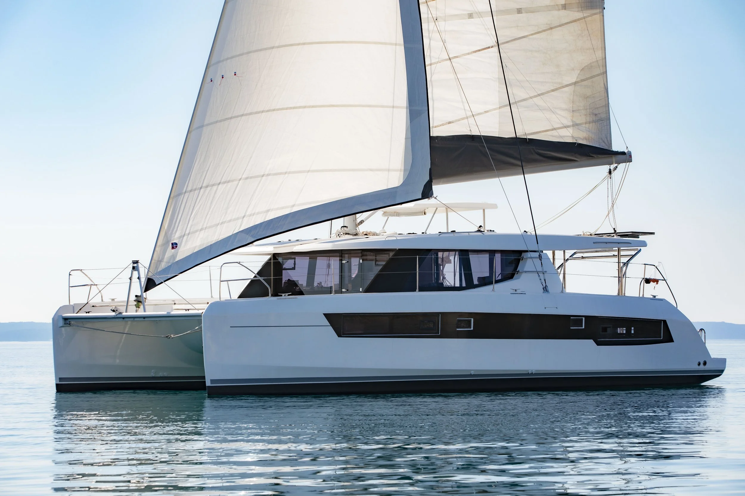 A modern white sailing catamaran with large sails on calm water under a clear sky.