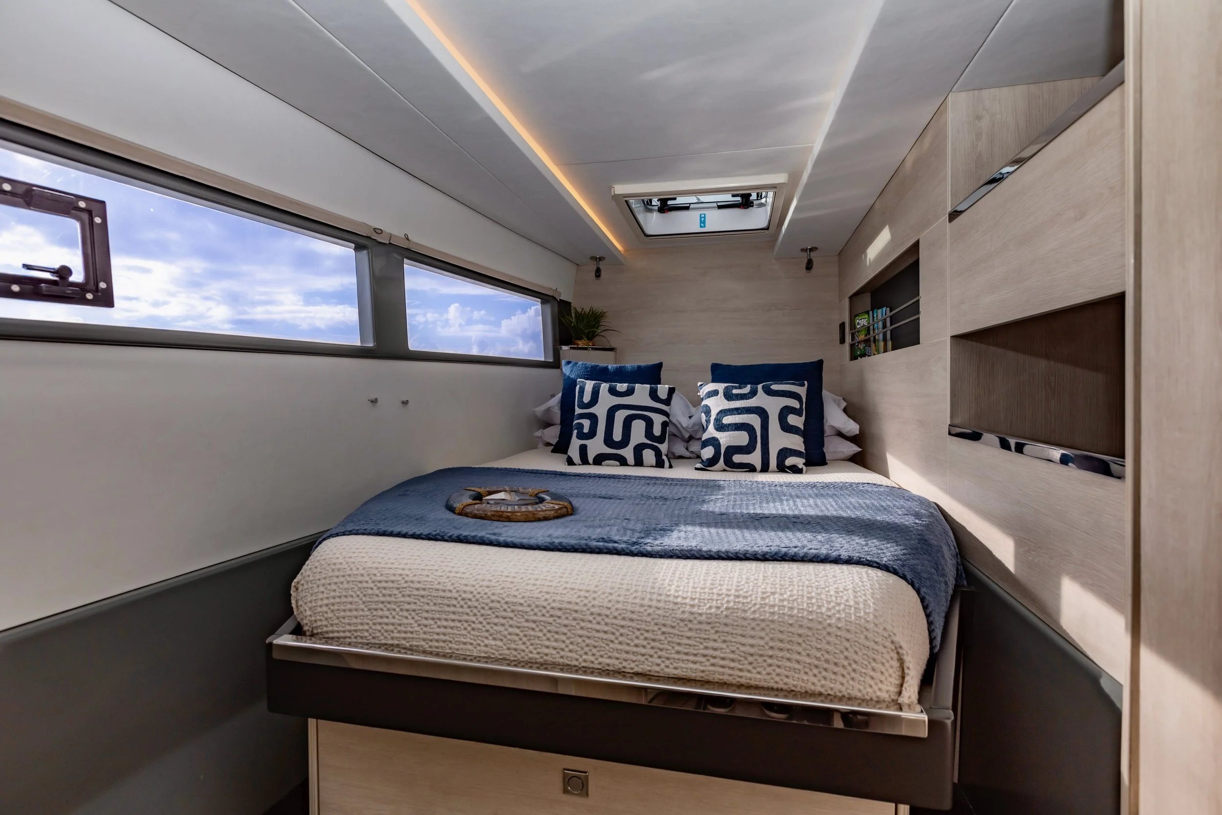 A cozy bedroom with a window view of the sky and clouds, a bed with blue and white pillows, a quilt, and a giraffe figurine on the bed, with built-in wooden shelves and a ceiling hatch.
