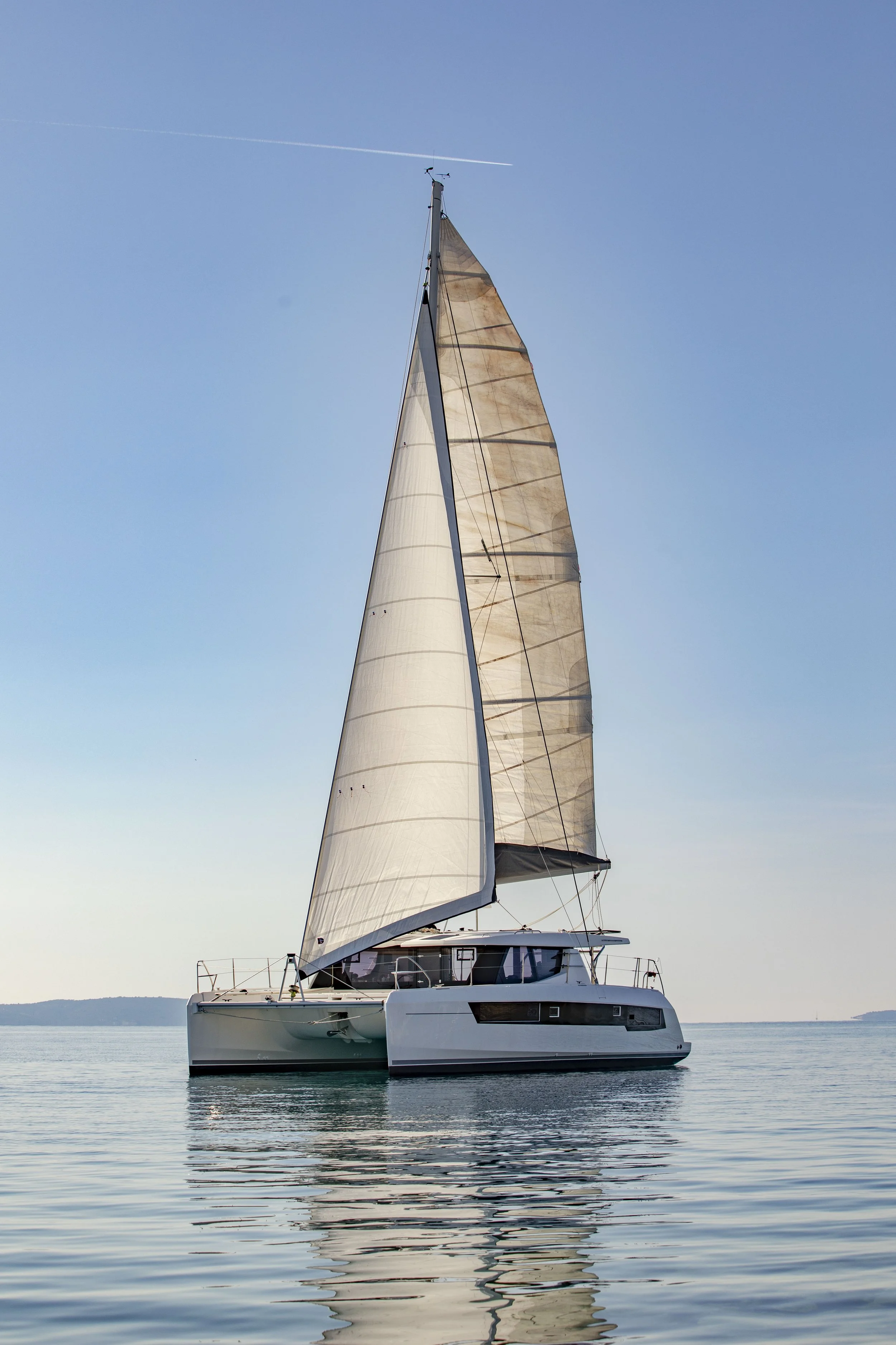 A modern sailboat with large beige sails floating on calm water under a clear blue sky.