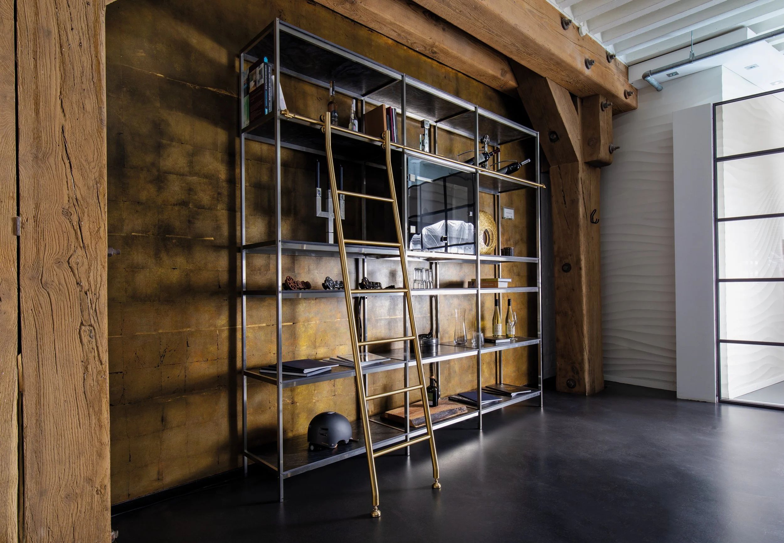 A modern interior with a metal shelving unit holding books, bottles, glasses, and decorative items. A gold-colored ladder leans against the shelf. The room has wooden beams, a dark floor, and white walls with a large window.