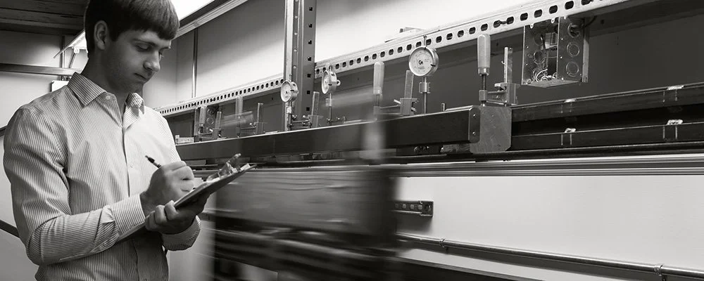A man in a striped shirt writing on a clipboard near a large industrial machine.