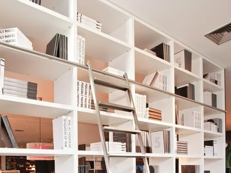 A white bookshelf with multiple compartments filled with books and decorative items, and a metal ladder leaning against it.