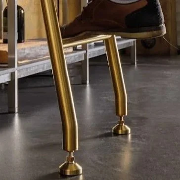 Close-up of a person's shoe resting on a shiny gold-colored chair leg with black rubber feet, on a dark floor near a metal bench.