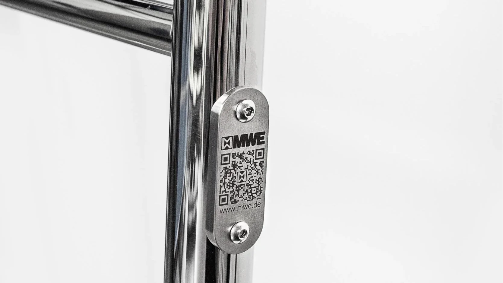 Close-up of a metallic door or panel with a silver tag labeled 'MWE,' QR code, and website 'www.mwe.de' attached with two screws. The background is plain and white.