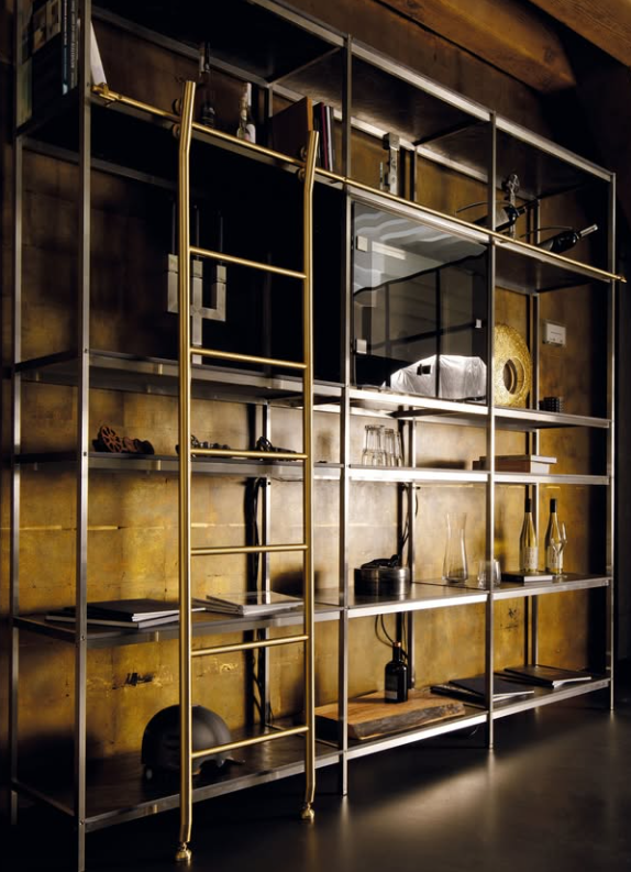 A tall metal and wood shelving unit with a brass ladder on the left side, filled with books, glasses, bottles, decorative objects, and books against a gold-toned wall.