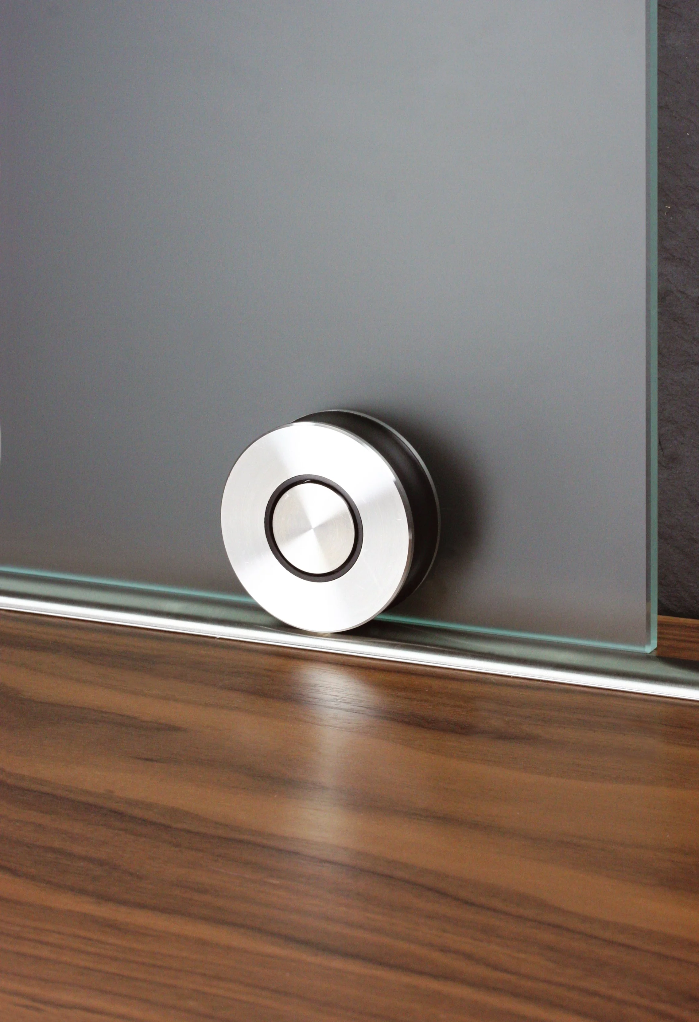 Close-up photograph of a modern metallic speaker with a silver and black circular dial, placed on a wooden surface next to a gray wall.