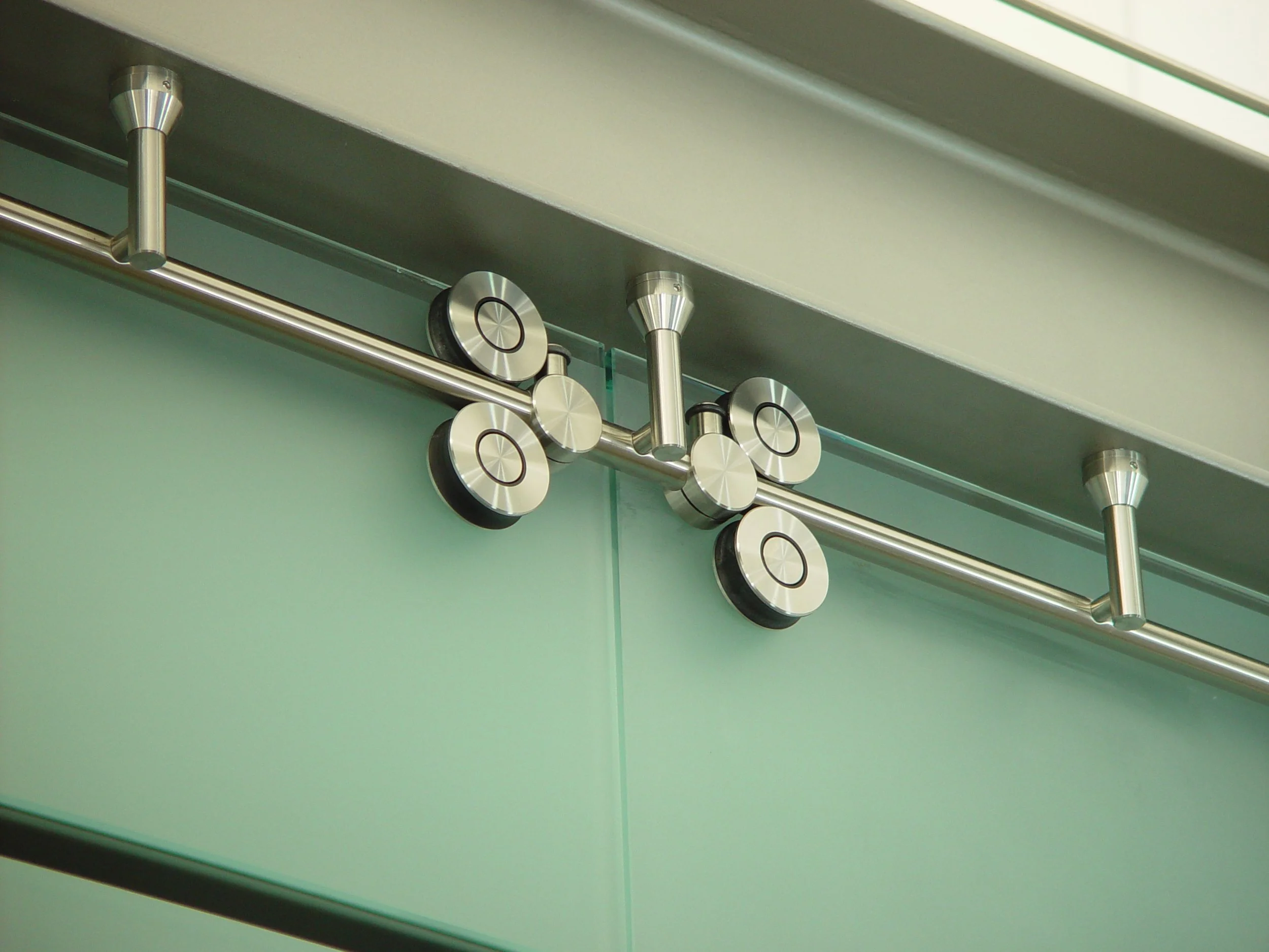 Close-up of a modern metallic door hinge with circular decorative elements, mounted on a glass door with a green background.