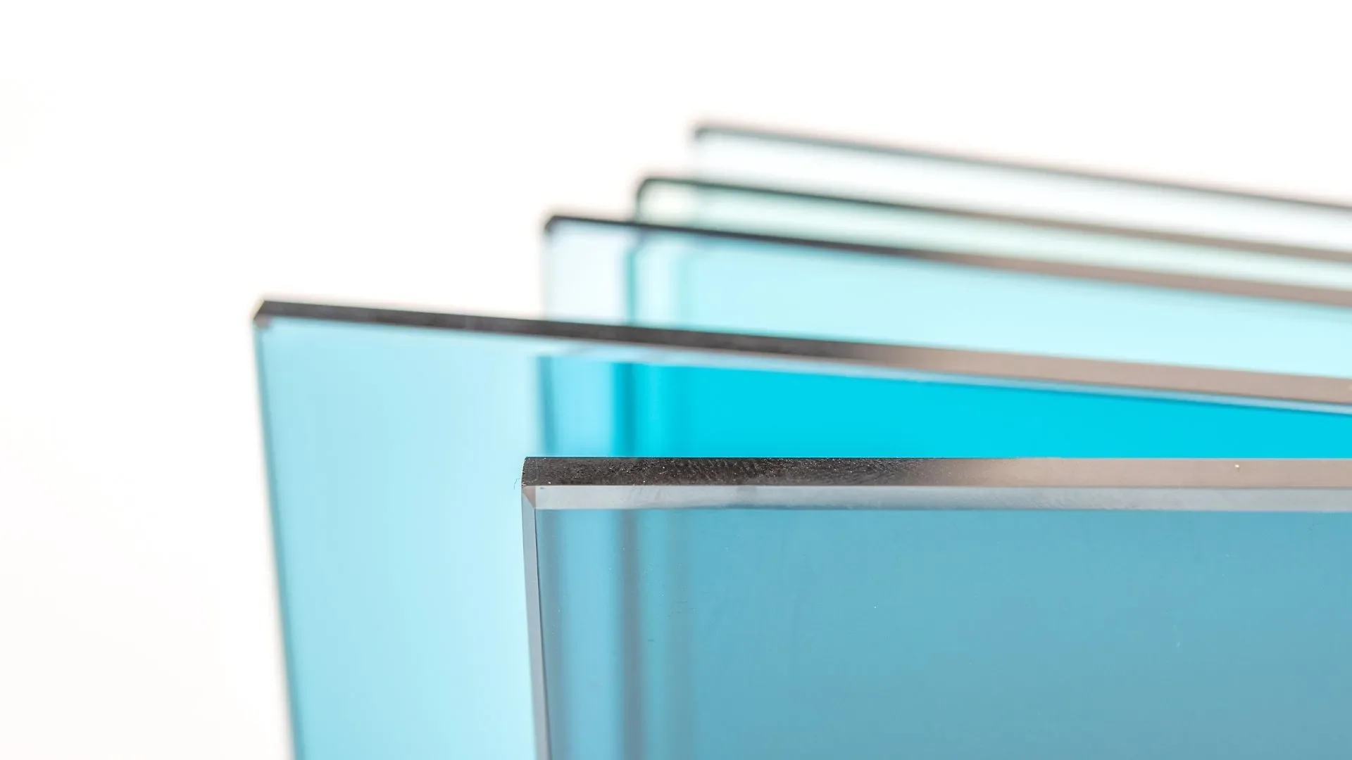 Close-up of several stacked sheet glass panels with blue-tinted transparent surfaces and metallic edges, arranged at an angle against a plain white background.