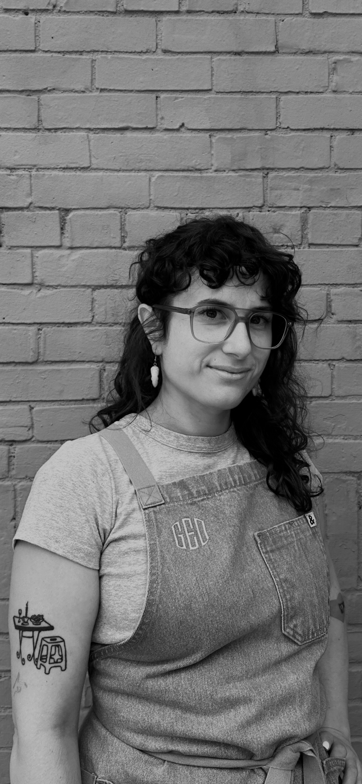 A woman with glasses and curly hair standing against a brick wall, wearing a t-shirt and denim apron with a tattoo on her arm.