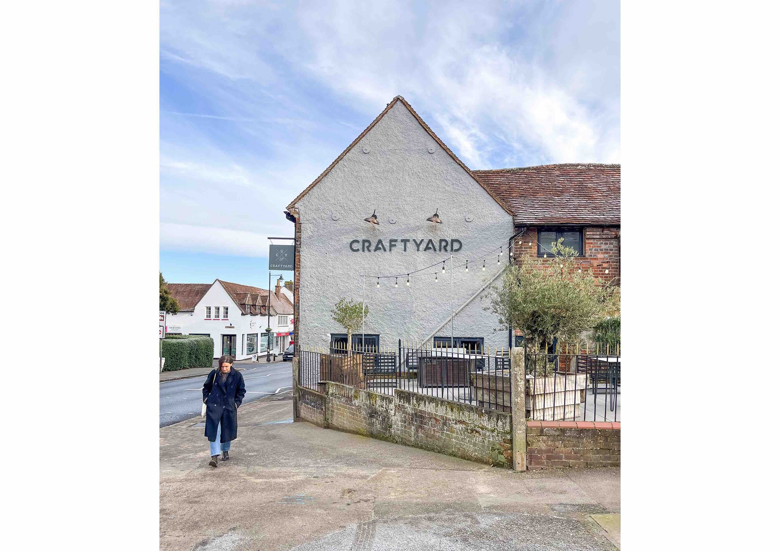 Exterior of building with "CRAFTYARD" sign, outdoor seating area, decorative lights, and a person walking along the sidewalk.