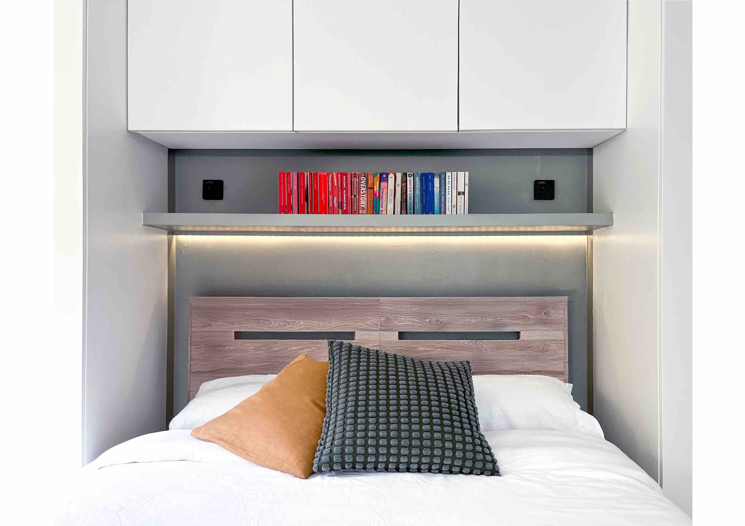 Modern bedroom with a wooden headboard, decorative cushions, floating shelf with books, under-cabinet lighting, and white cabinets above.