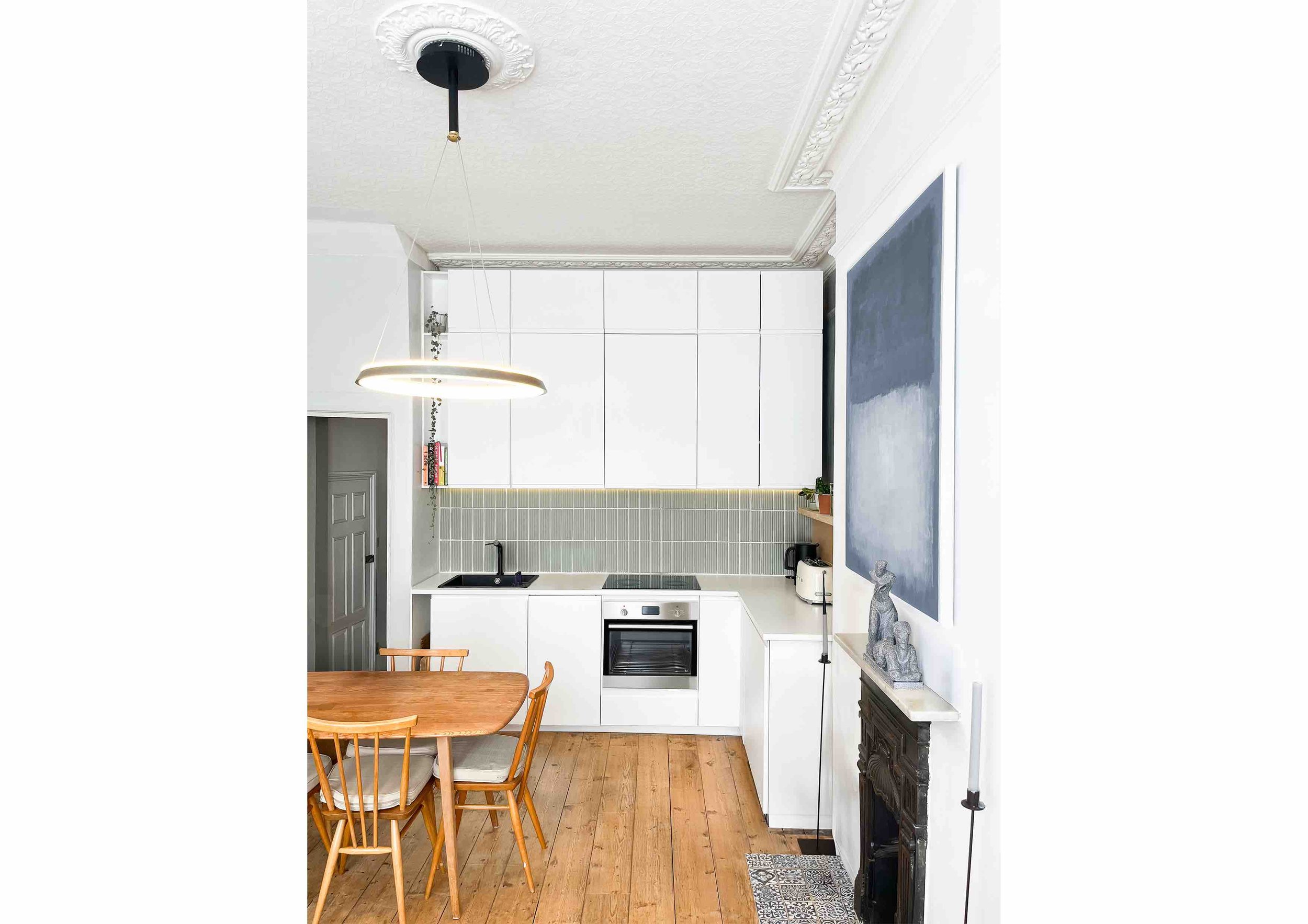 Modern kitchen with white cabinets, wood dining table, and circular pendant light. Features include a black sink, built-in oven, tiled backsplash, and decorative wall art.