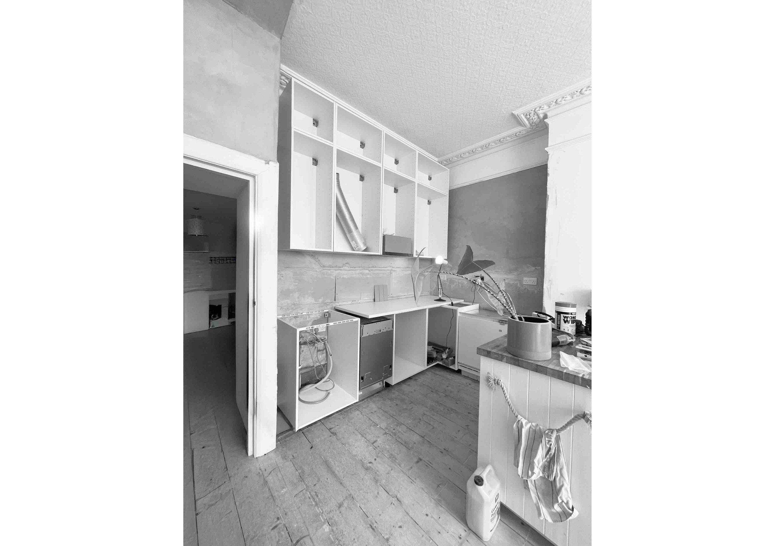 Monochrome photo of a kitchen under renovation with open cabinets, tools, and a lamp.