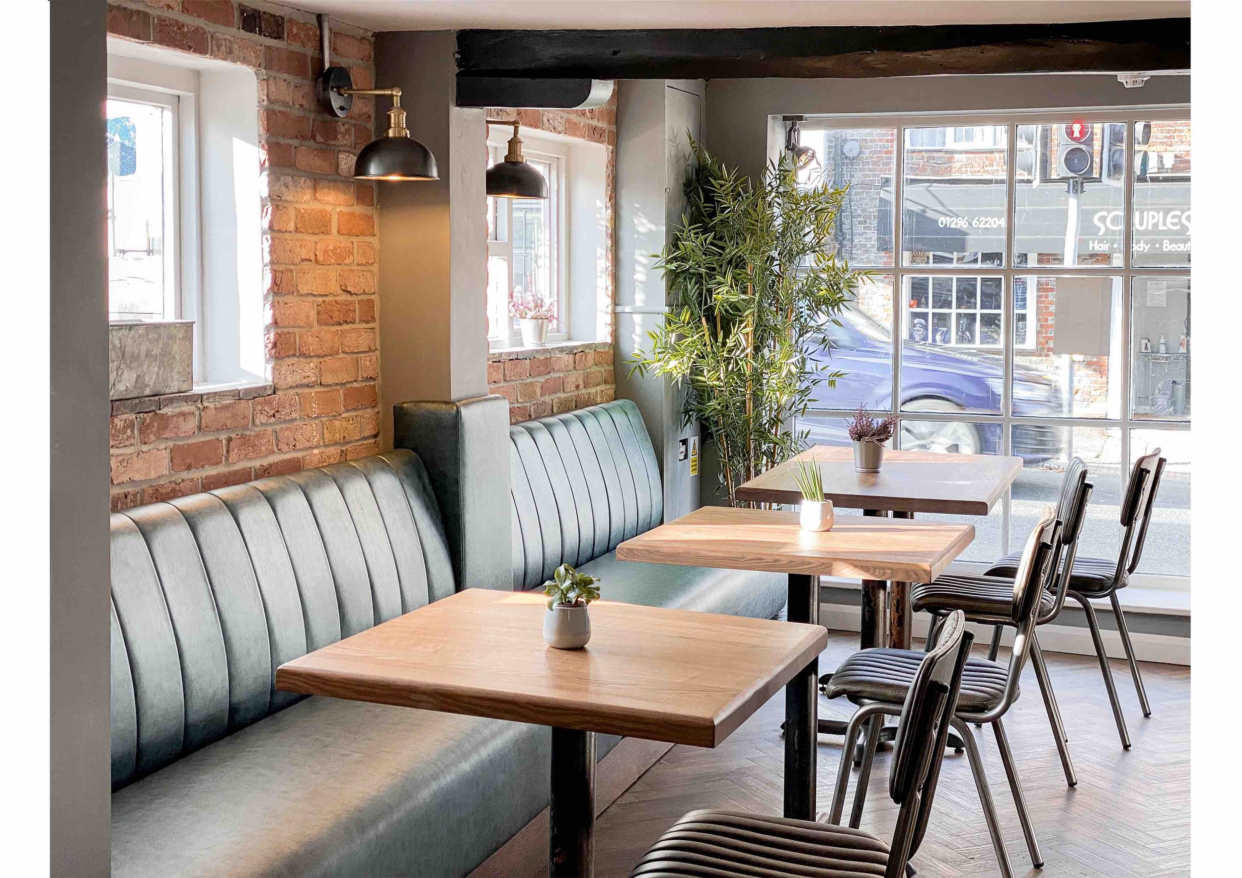 Modern cafe interior with brick walls, padded seating, wooden tables, chairs, potted plants, and large windows.