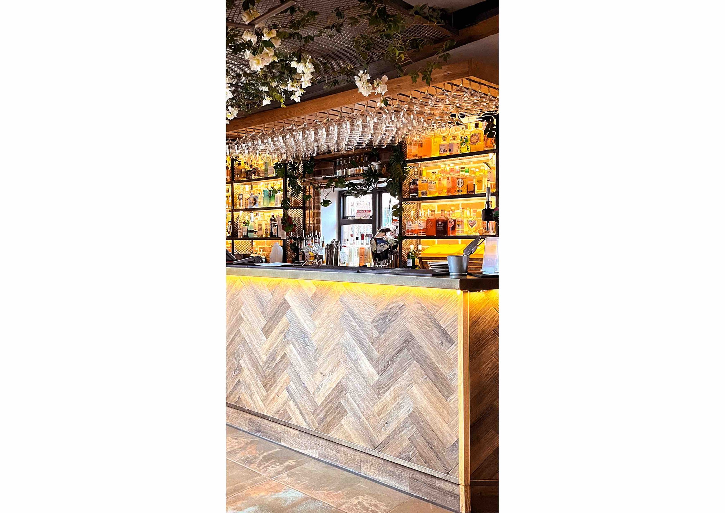 Modern bar interior with wood paneling, hanging wine glasses, illuminated shelves with bottles, and decorative greenery.