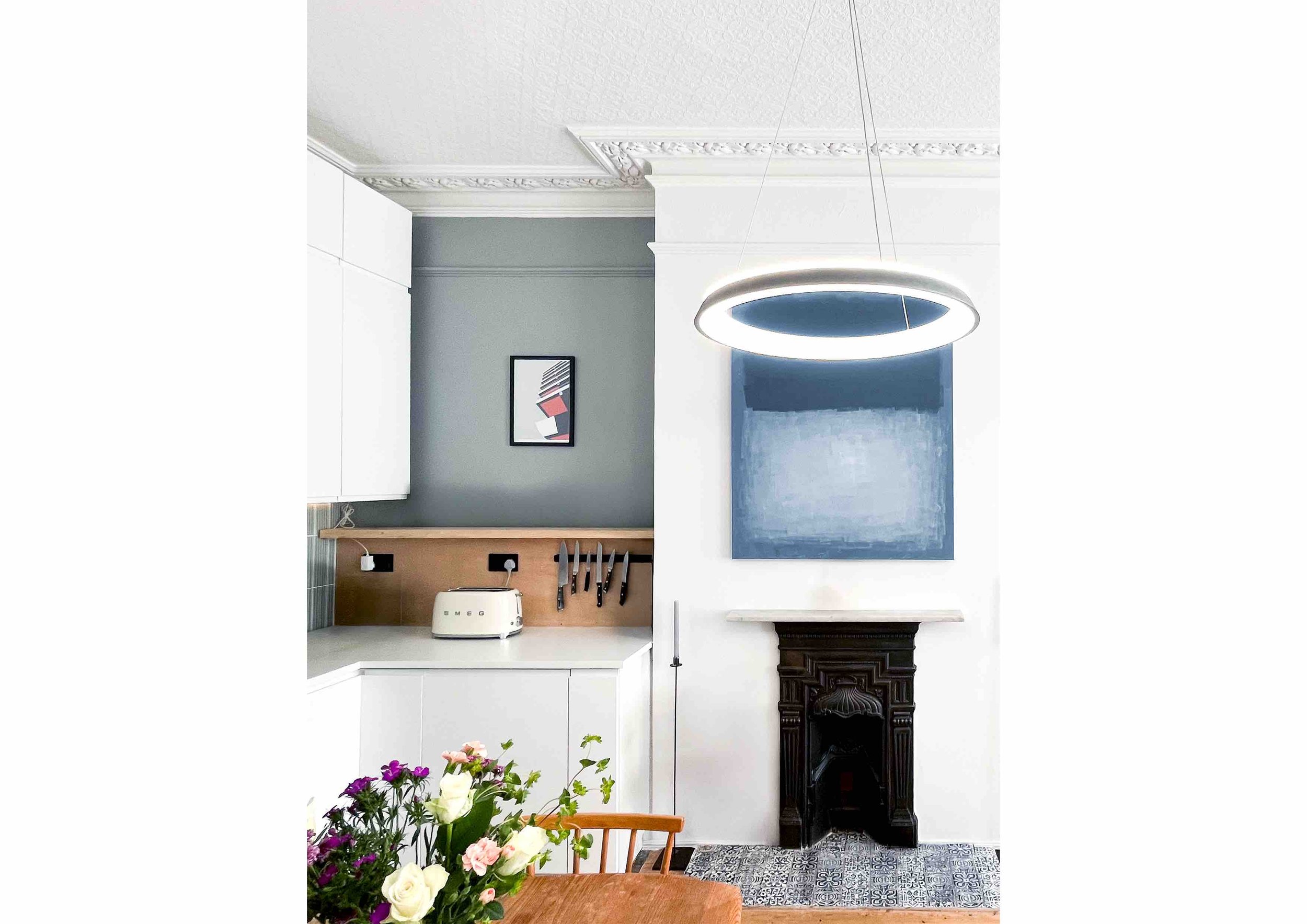 Modern kitchen with white cabinets, Smeg toaster, hanging knives, abstract art, floral arrangement, and decorative ceiling.