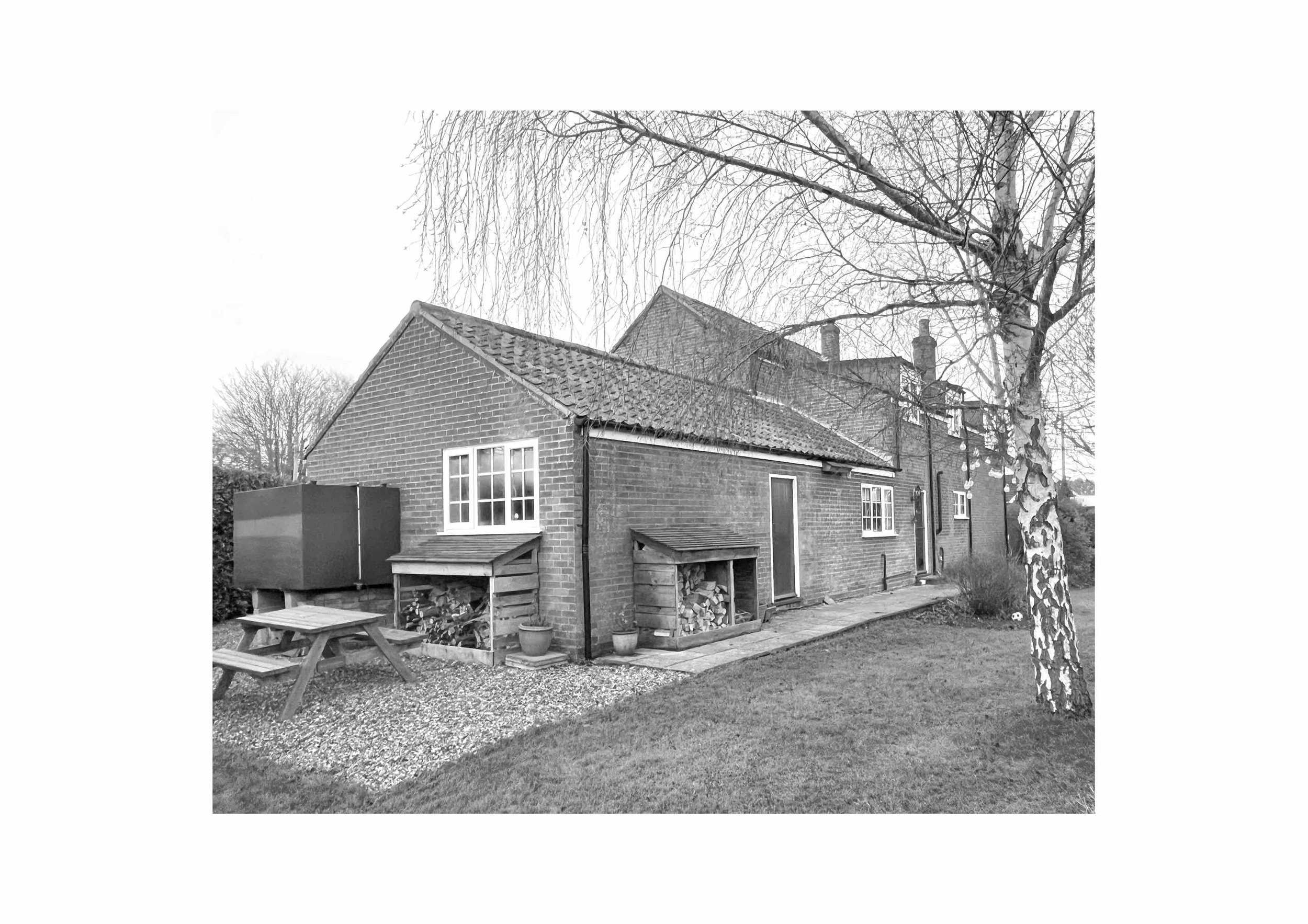 Black and white photo of a brick house with tiled roof, wood storage sheds, and a picnic table. A birch tree is in the foreground.