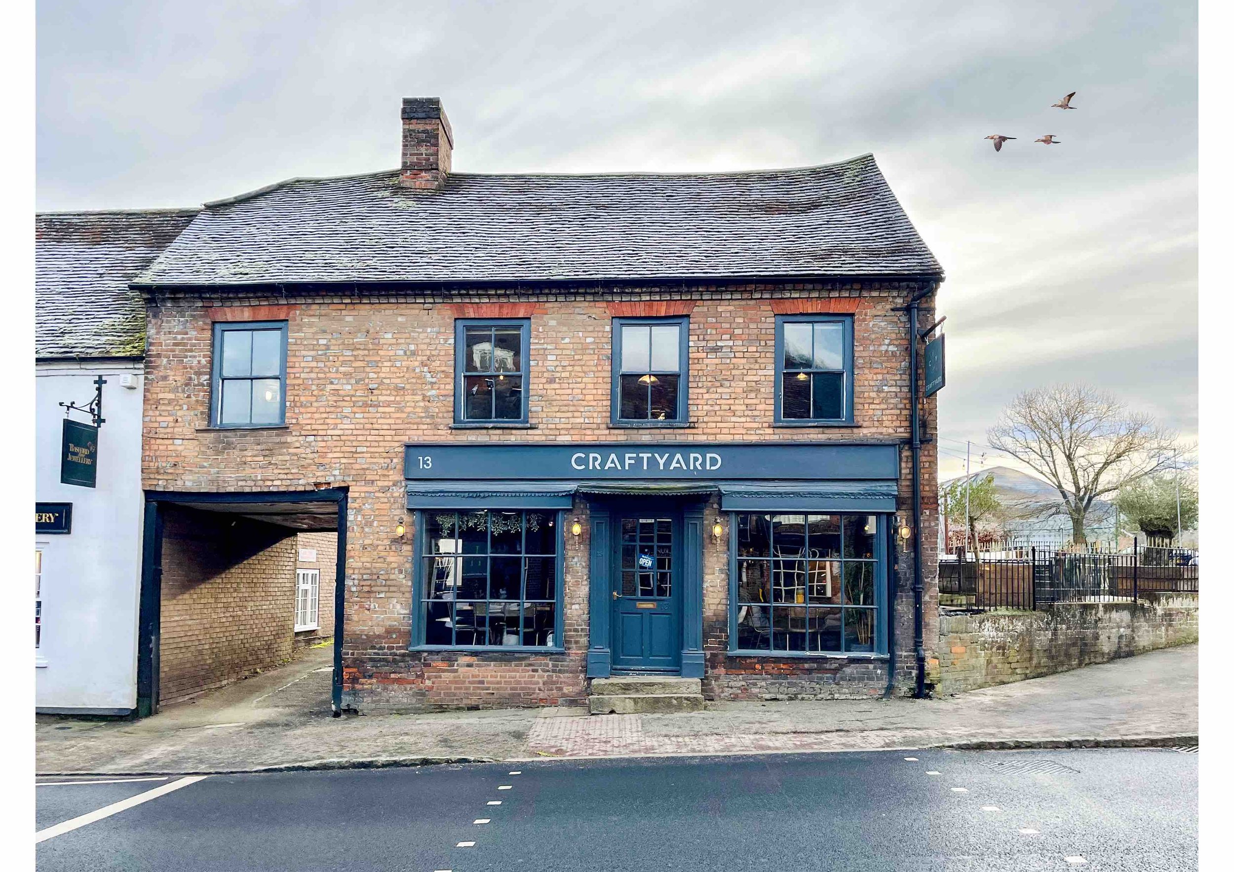 Exterior of a brick building with blue signage labeled 'CRAFTYARD', featuring three windows and a central door, set on a street with birds flying in the sky.