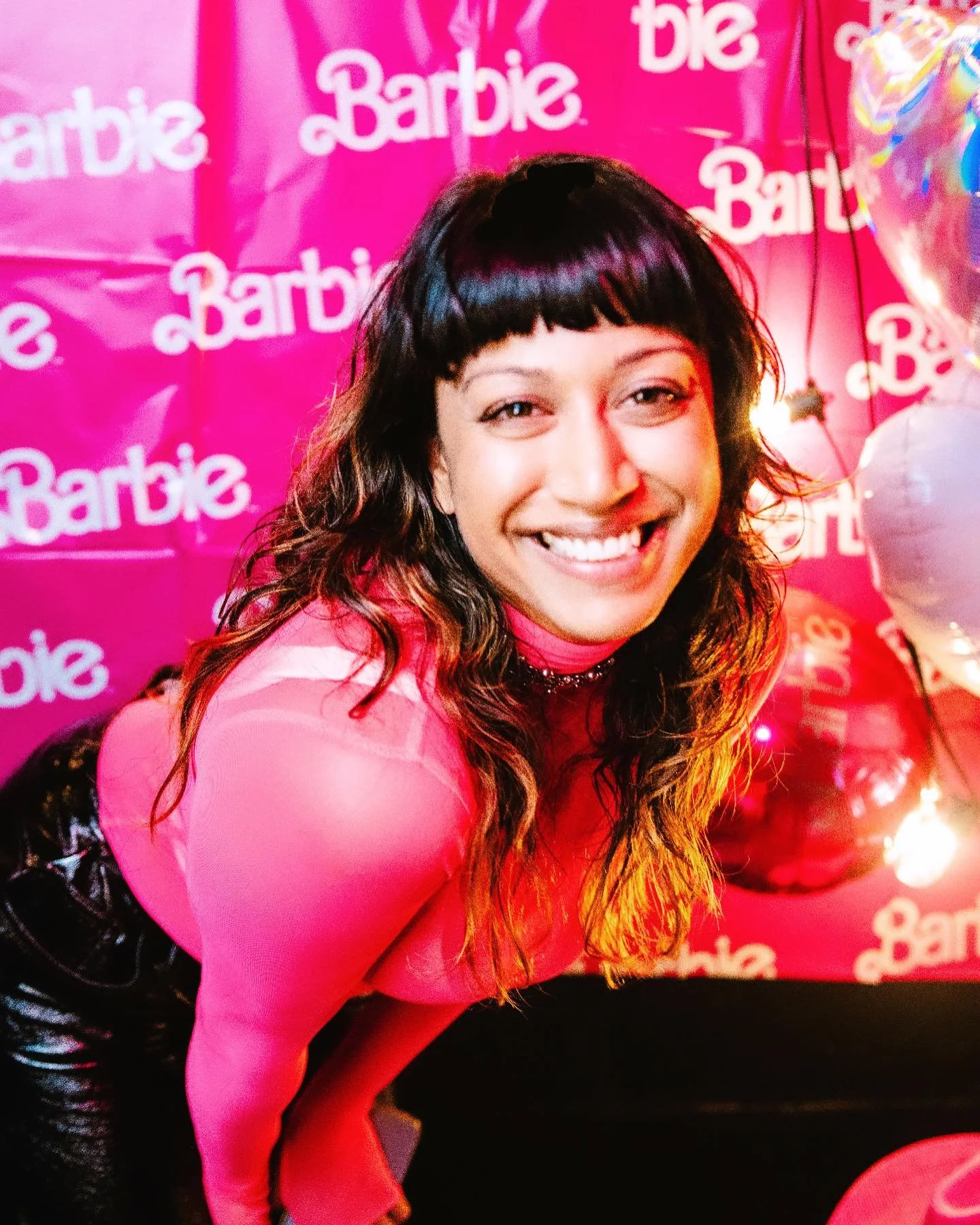 Person smiling in front of a pink Barbie-themed backdrop, wearing a pink top and surrounded by balloons.