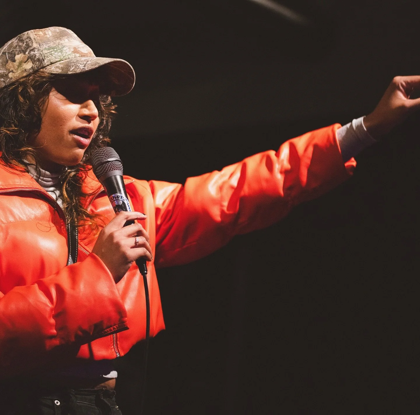 Person in an orange jacket and camouflage cap holding a microphone, gesturing with one arm raised.