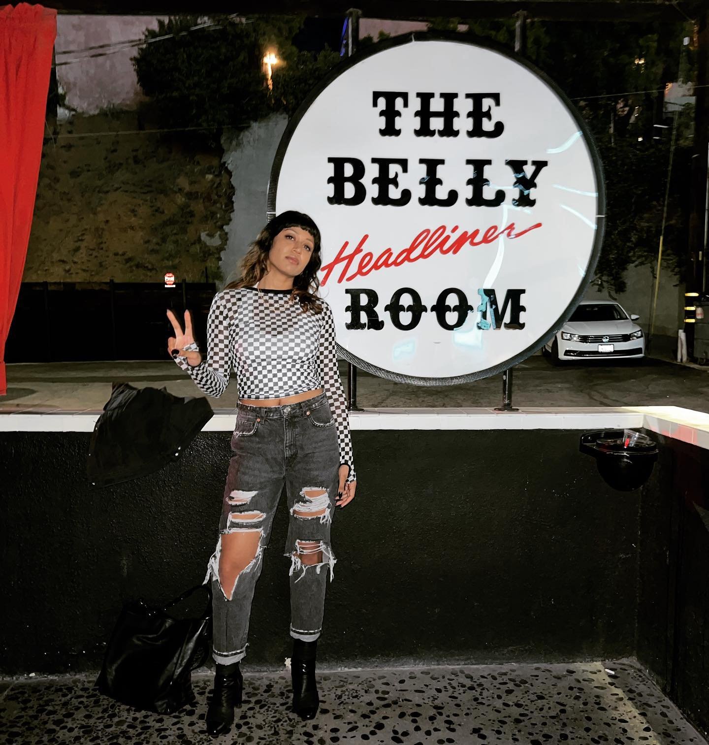 Person in checkered crop top and ripped jeans posing with a peace sign next to 'The Belly Room' sign.