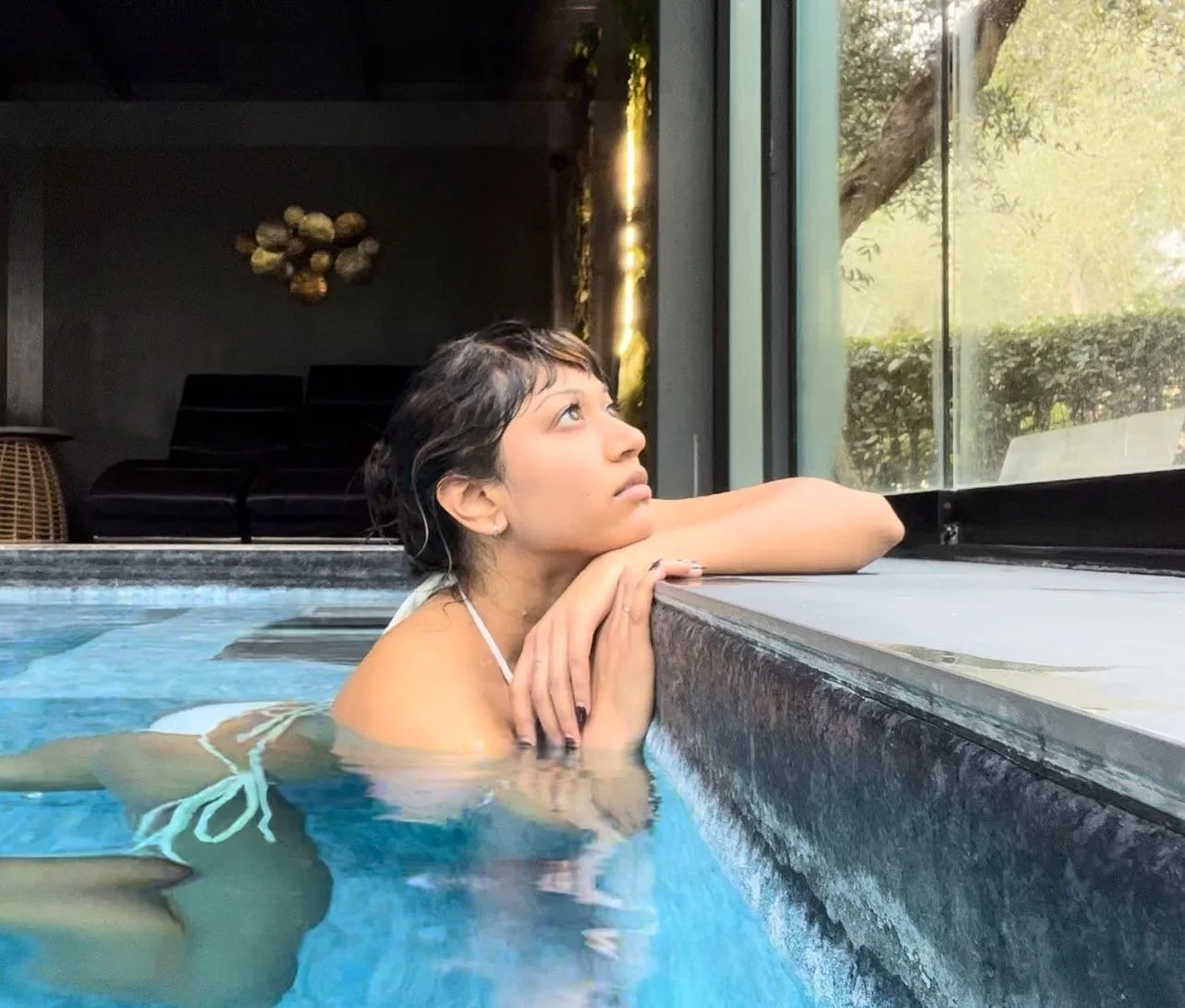 Person in a bikini leaning on the edge of an indoor pool, gazing out a large window.