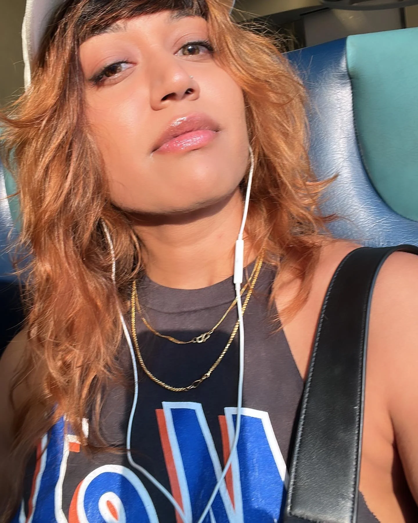 A woman with curly hair wearing a cap, sleeveless top, and earphones, sitting on a train seat.