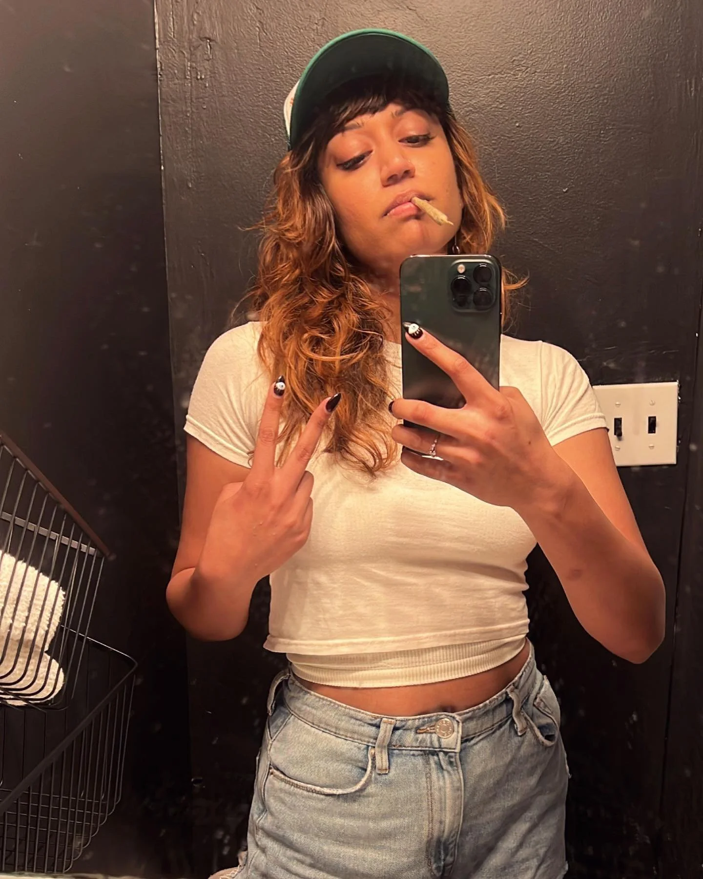 A person taking a selfie in a mirror, wearing a cap, white cropped t-shirt, and denim shorts, holding up a peace sign and holding a phone.