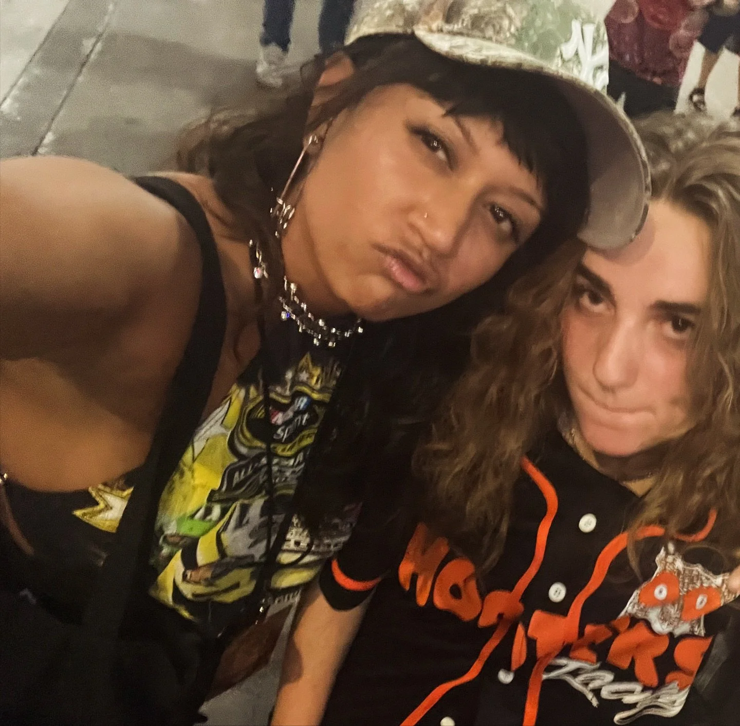 Two people taking a selfie, one wearing a baseball cap and graphic shirt, the other in a Hooters jersey. Street setting.