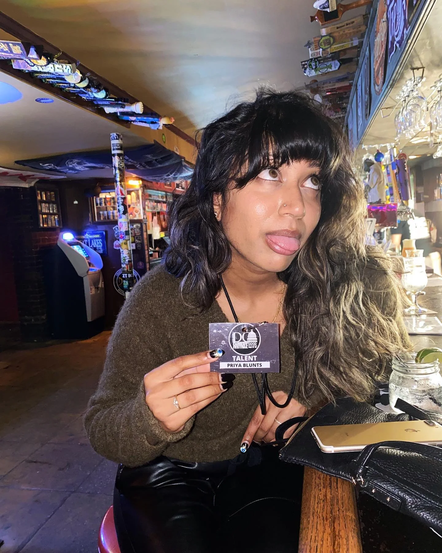Person sitting at a bar holding a talent pass, sticking out their tongue, with long dark hair and wearing a dark sweater.