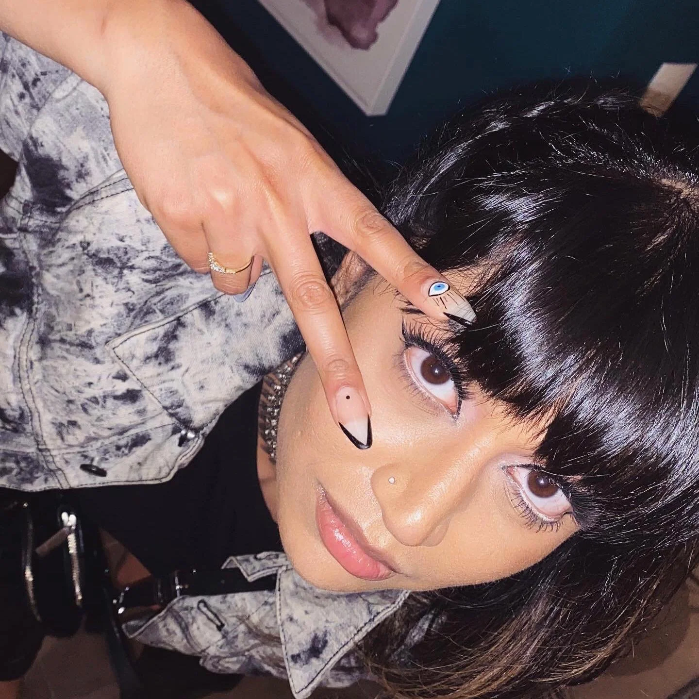 Person with bangs and patterned nails making a peace sign gesture close to their eye.