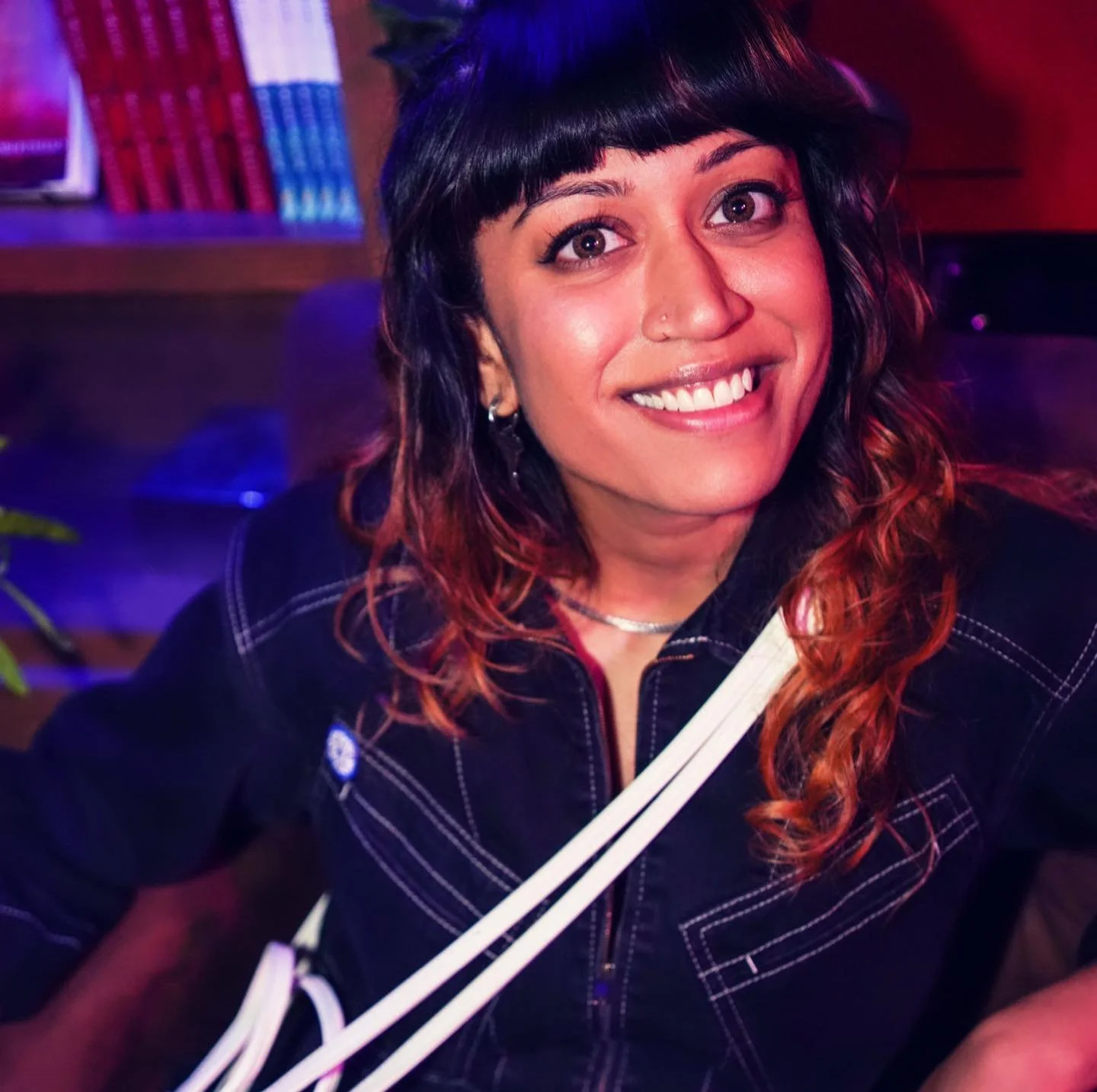 Smiling person with dark hair and a nose ring, wearing a black top, sitting in a dimly lit room with blurred bookshelves in the background.