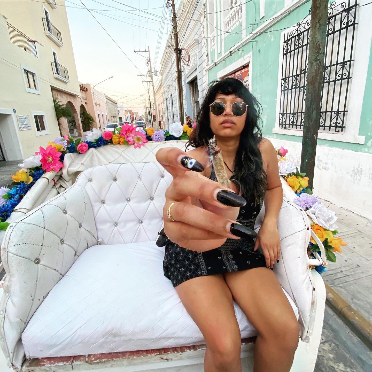 Person sitting on a white, tufted seat decorated with colorful flowers, offering a hand forward with black nail polish and long nails. They are wearing sunglasses and sitting in an outdoor urban setting.