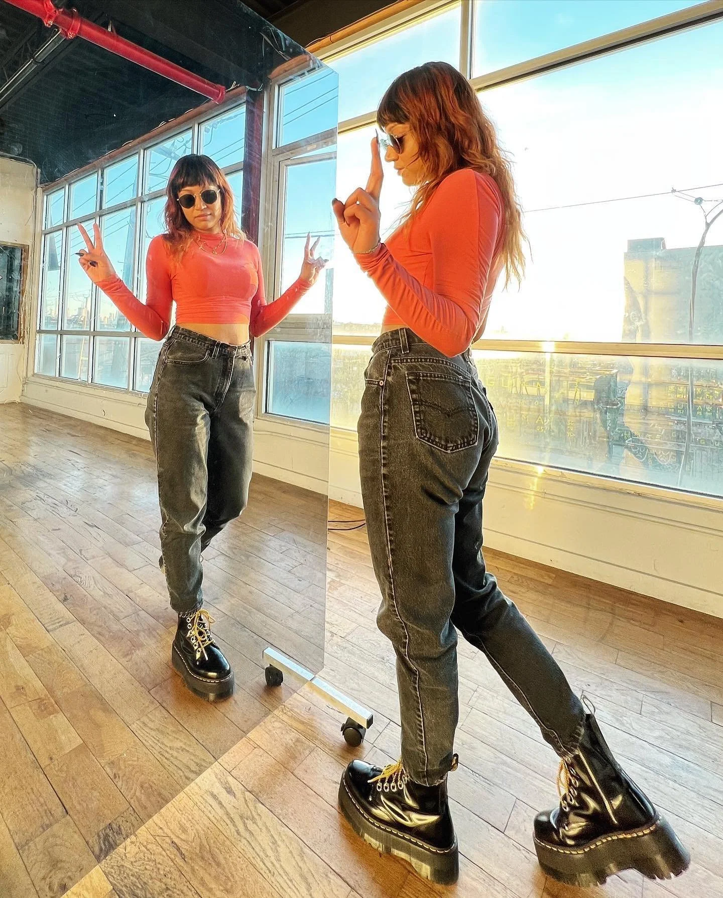 Woman in orange top and jeans posing in front of a mirror inside a sunlit room with wooden floors, wearing sunglasses and black boots.