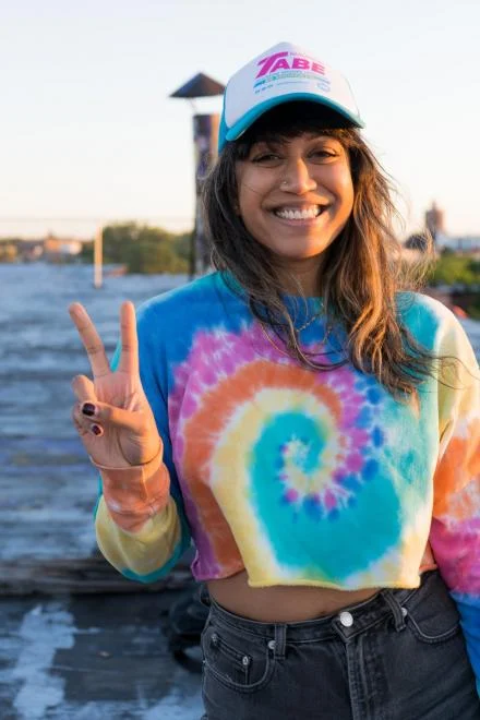 Smiling person wearing a colorful tie-dye crop top and a white cap, making a peace sign with their fingers, standing near water.