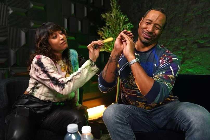 Two people sitting indoors, one holding a popsicle, the other playfully reacting with a cringe expression. The background has a plant and textured walls. Bottles of water are on the table in the foreground.