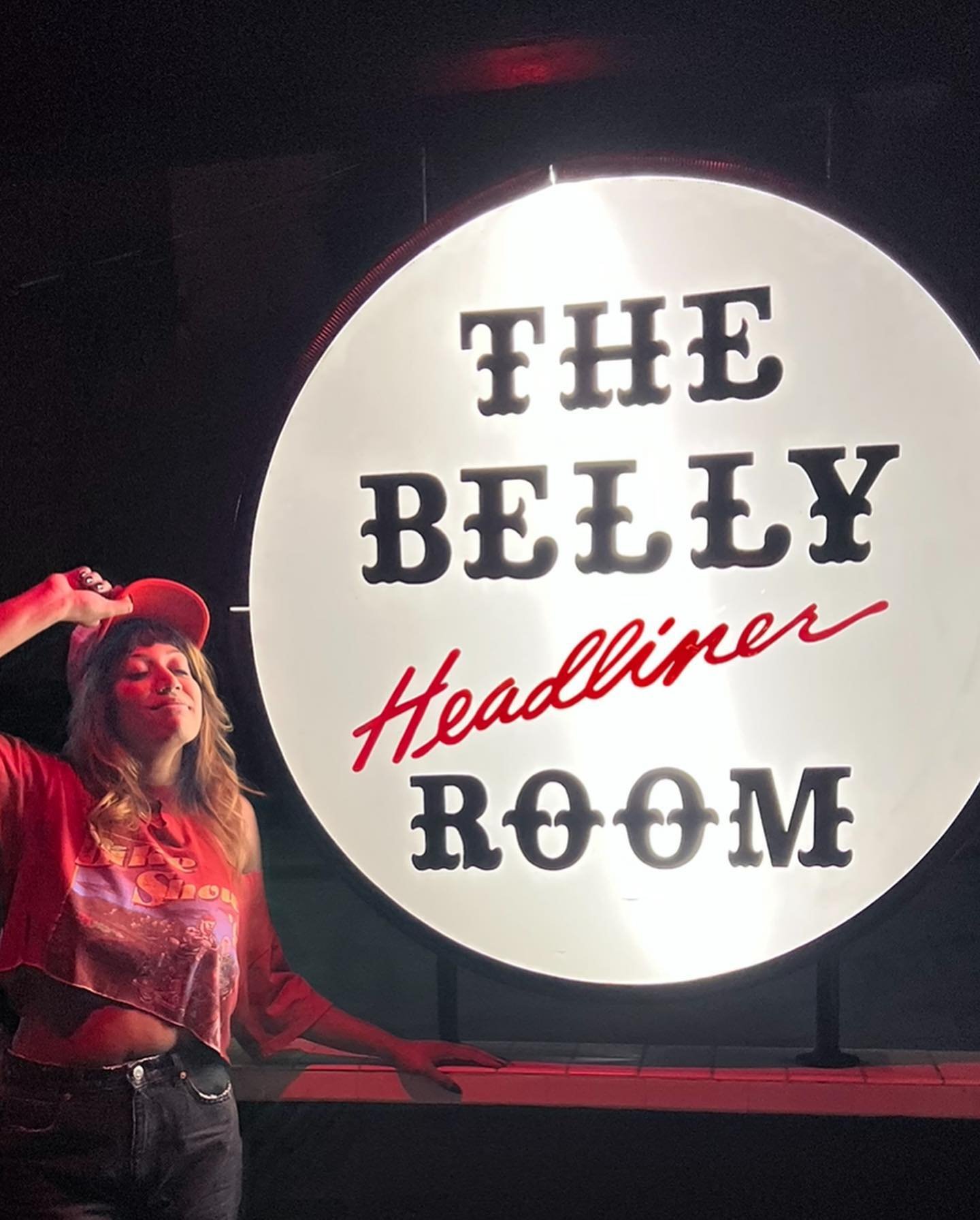 Person standing next to a circular sign illuminated with the text "The Belly Room Headliner," wearing a red cap and casual outfit in a dimly lit setting.